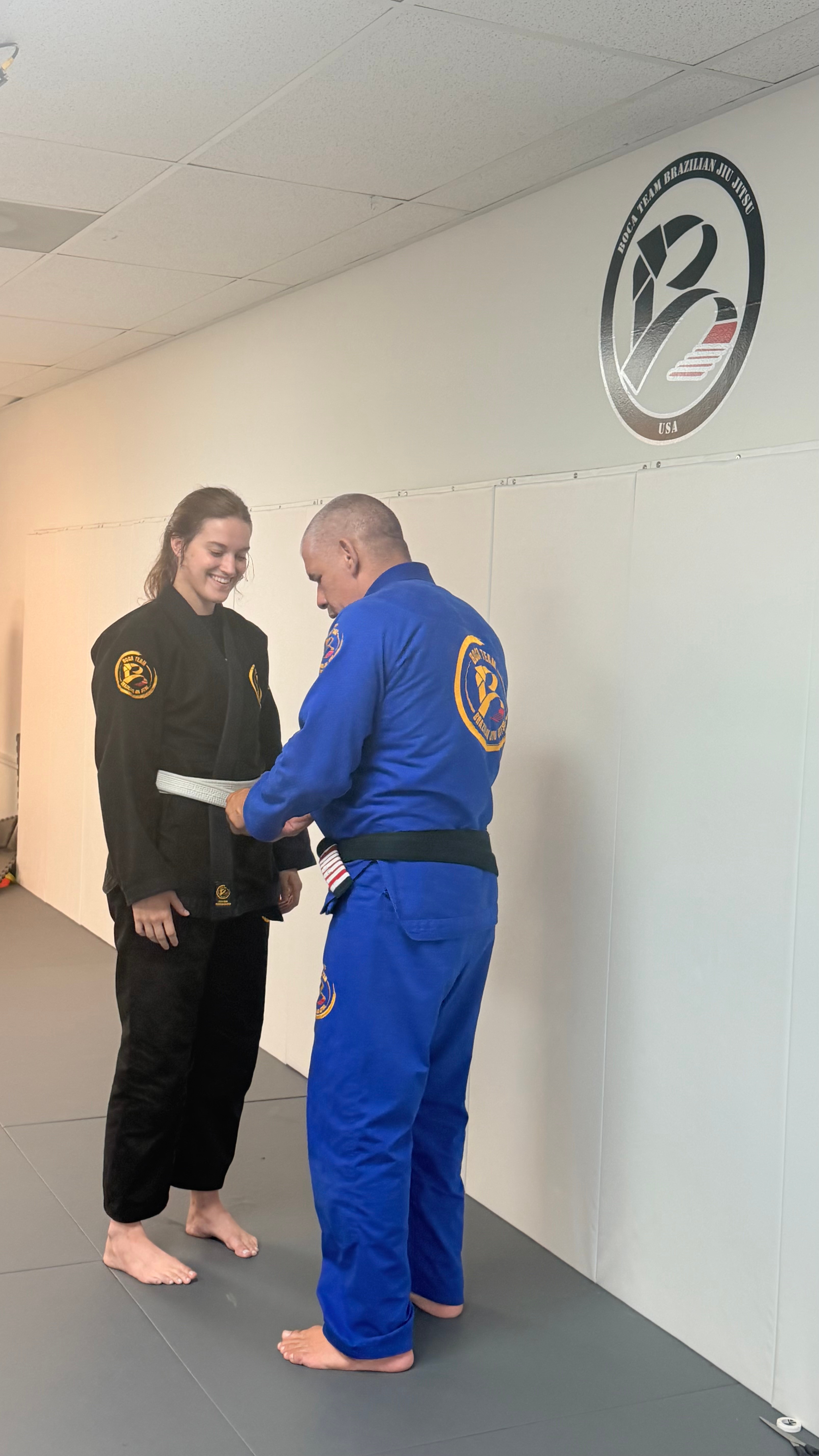 Two people in martial arts uniforms exchanging a certificate in a training room, with a logo on the wall.