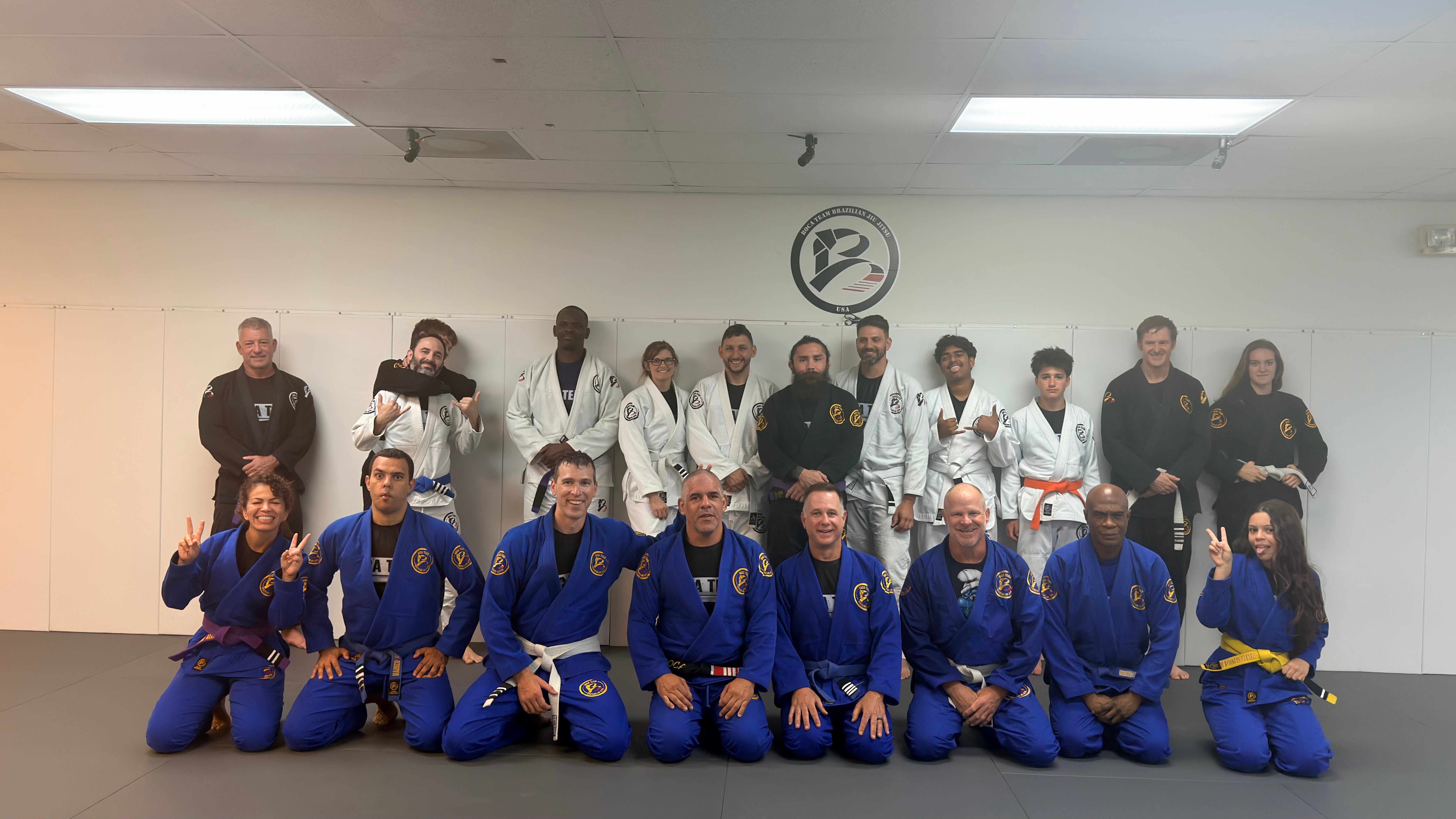 Group of people in martial arts uniforms posing indoors, some kneeling and some standing, with a clock on the wall behind them.