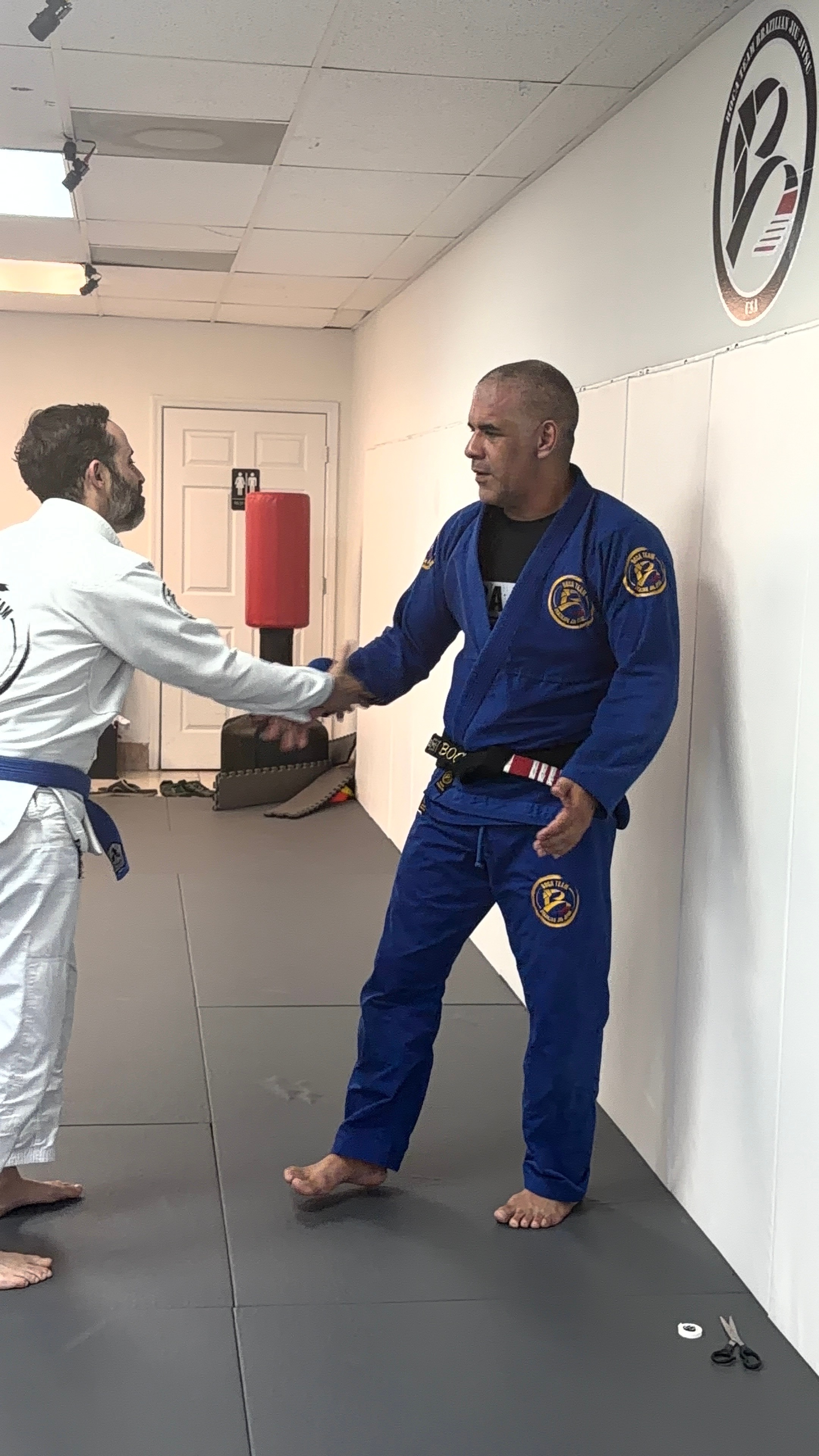 Two people shaking hands in a martial arts training room, one in a white gi and the other in a blue gi, with a white wall and logo behind them.