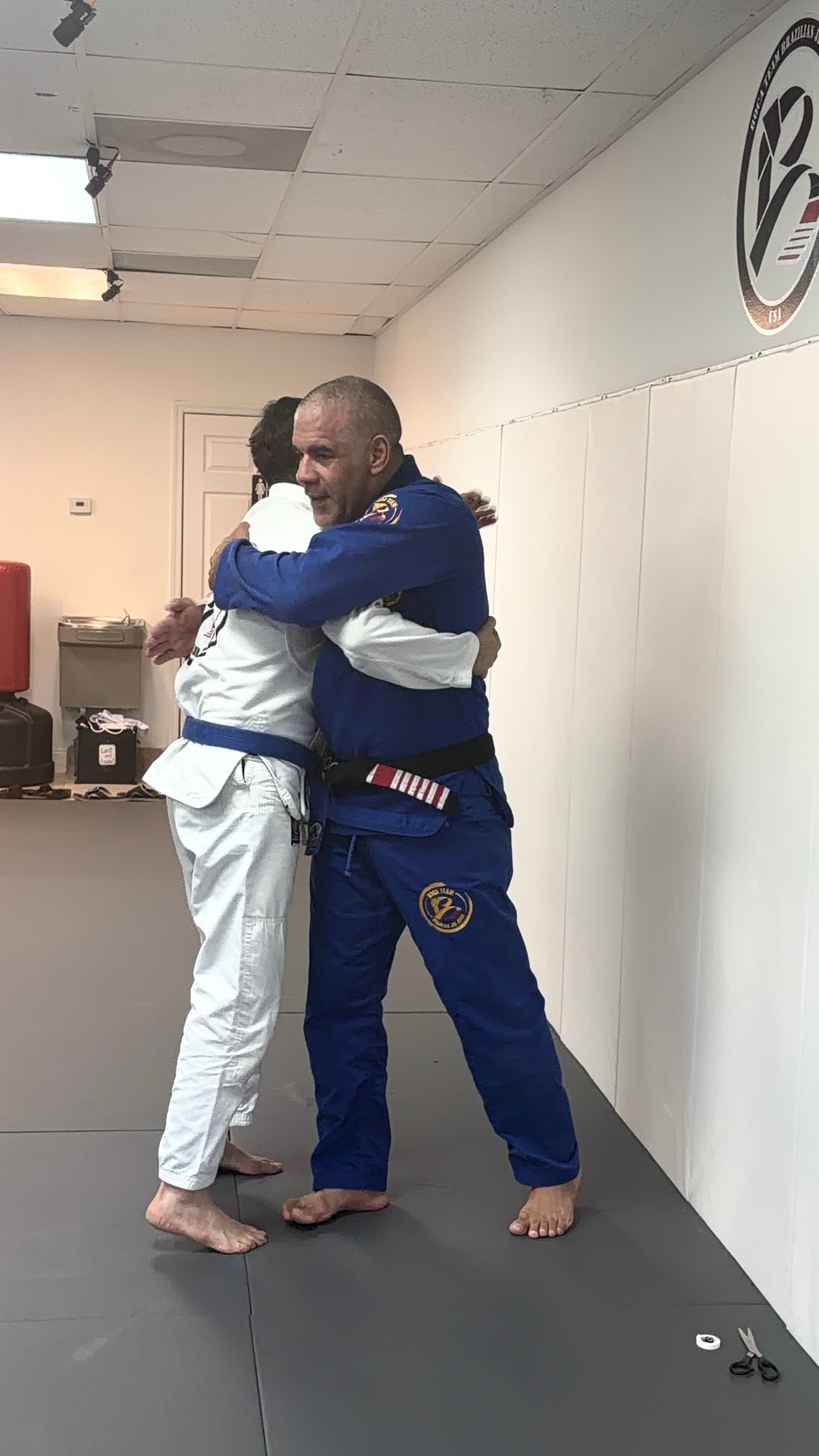 Two people in martial arts uniforms hugging in a training room, one in white and one in blue, with a white wall and logo in background.