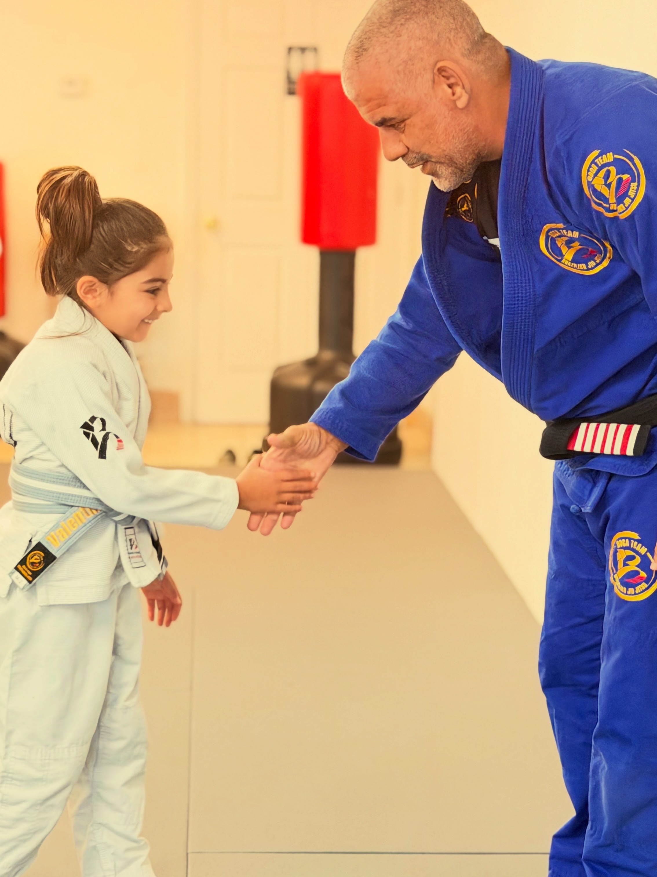 A young girl in a martial arts uniform shaking hands with an adult in a blue martial arts gi, indoors.