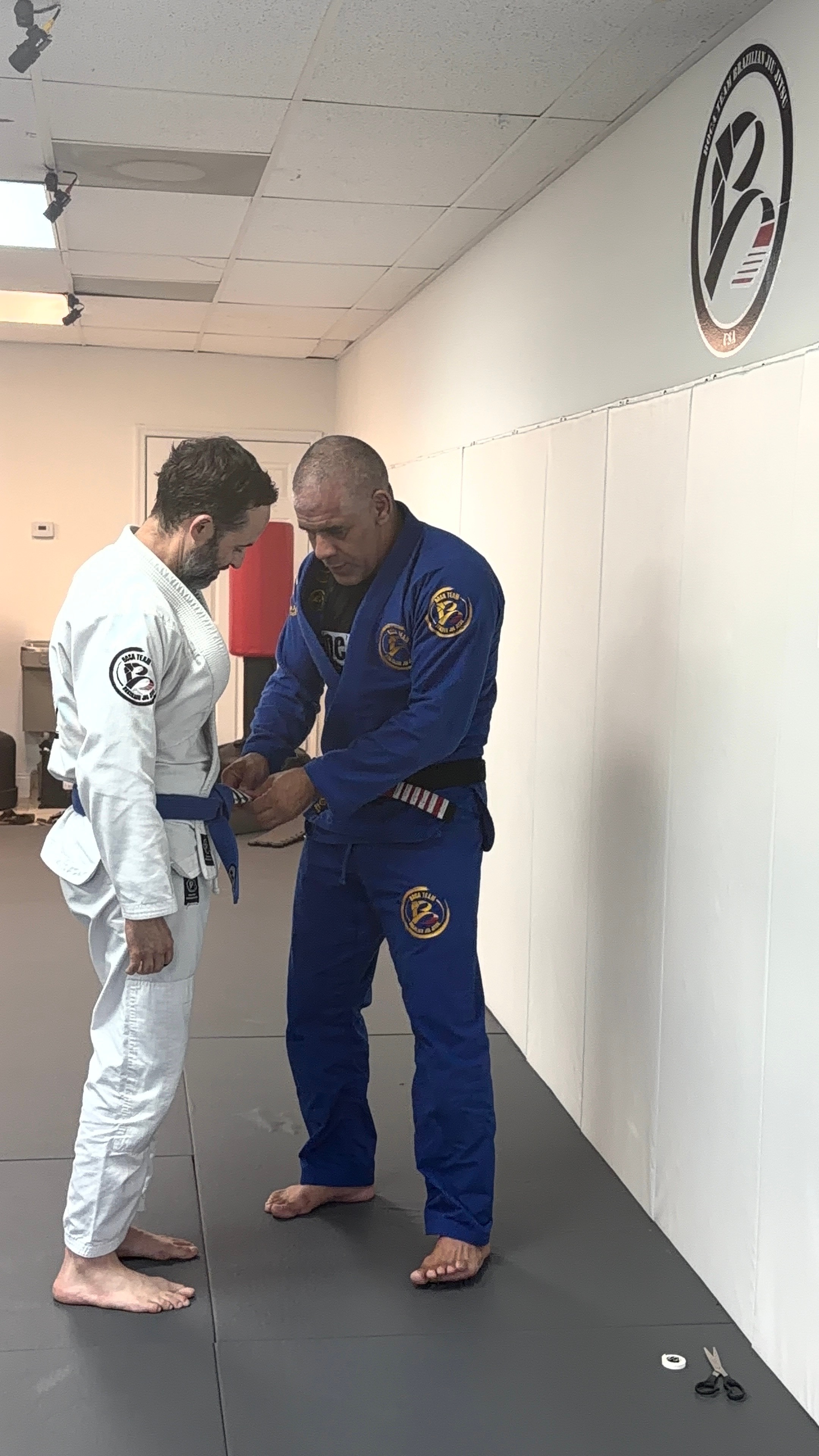 Two people in Brazilian Jiu-Jitsu uniforms, one in white and one in blue, standing on a mat, shaking hands.
