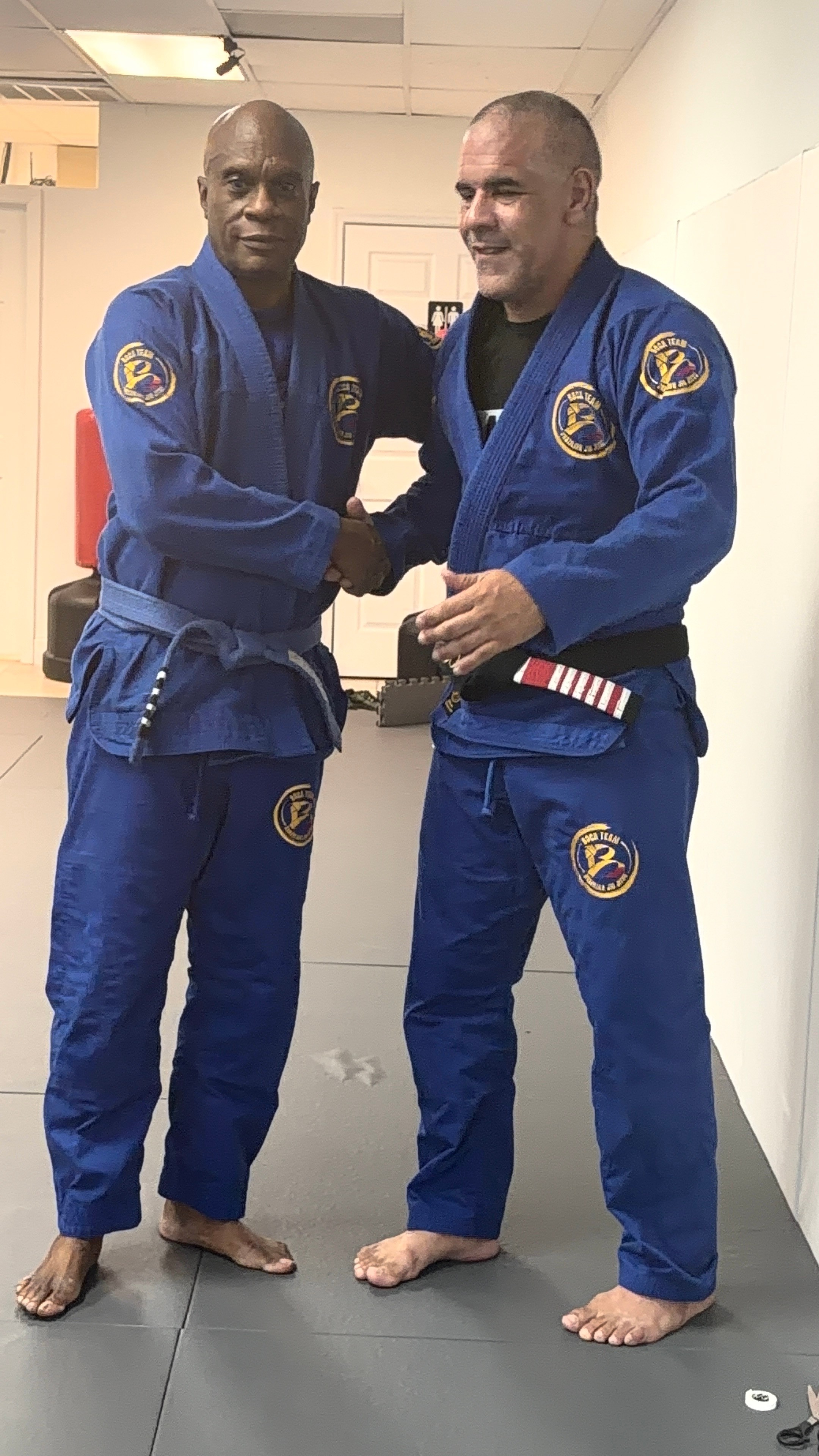 Two men in blue martial arts uniforms shaking hands, standing on a gray mat in a training room.