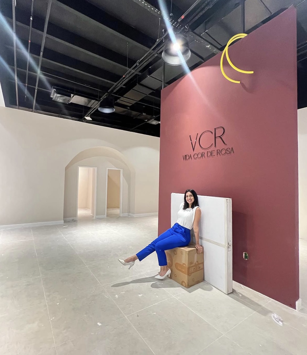 Woman sitting on a wooden box against a pink wall with text, in a spacious, modern interior with high ceilings.