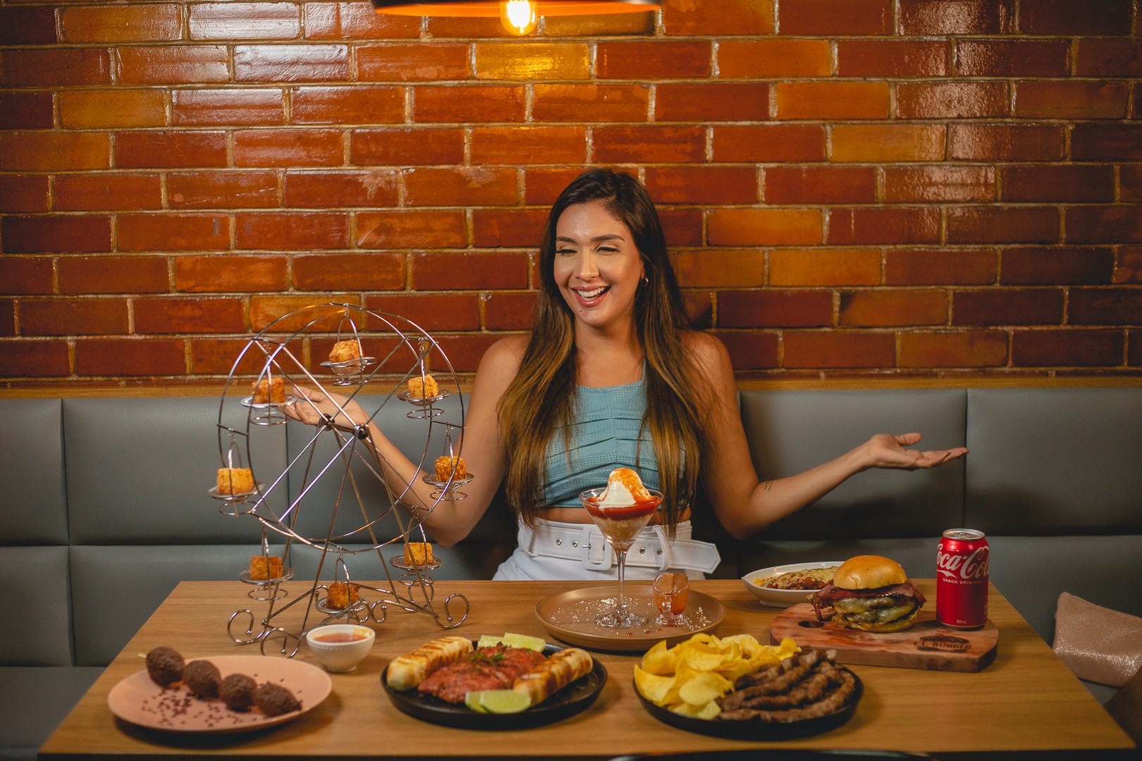 Smiling woman with long hair sitting at a table with food and a Ferris wheel dessert display, brick wall background.