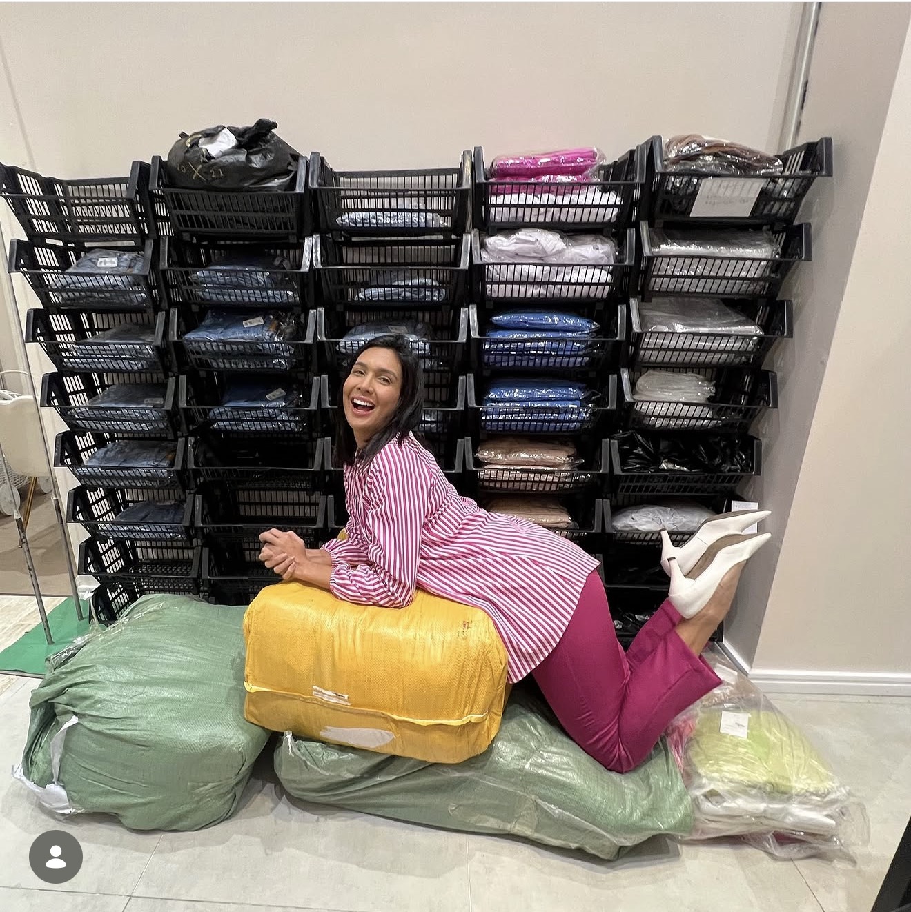 Person lying on large bags, smiling, in front of stacked black baskets, wearing pink and yellow clothing.