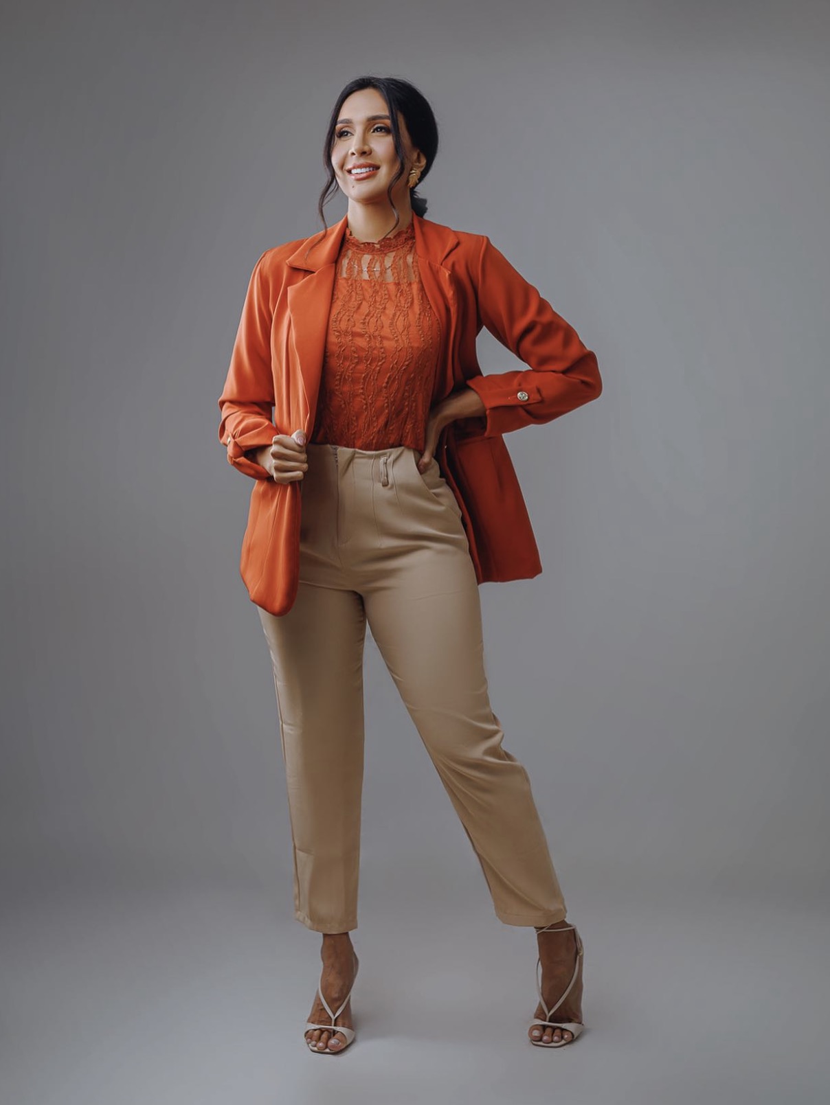 Woman in orange blazer and beige pants smiling, standing with hands on hips against gray background.
