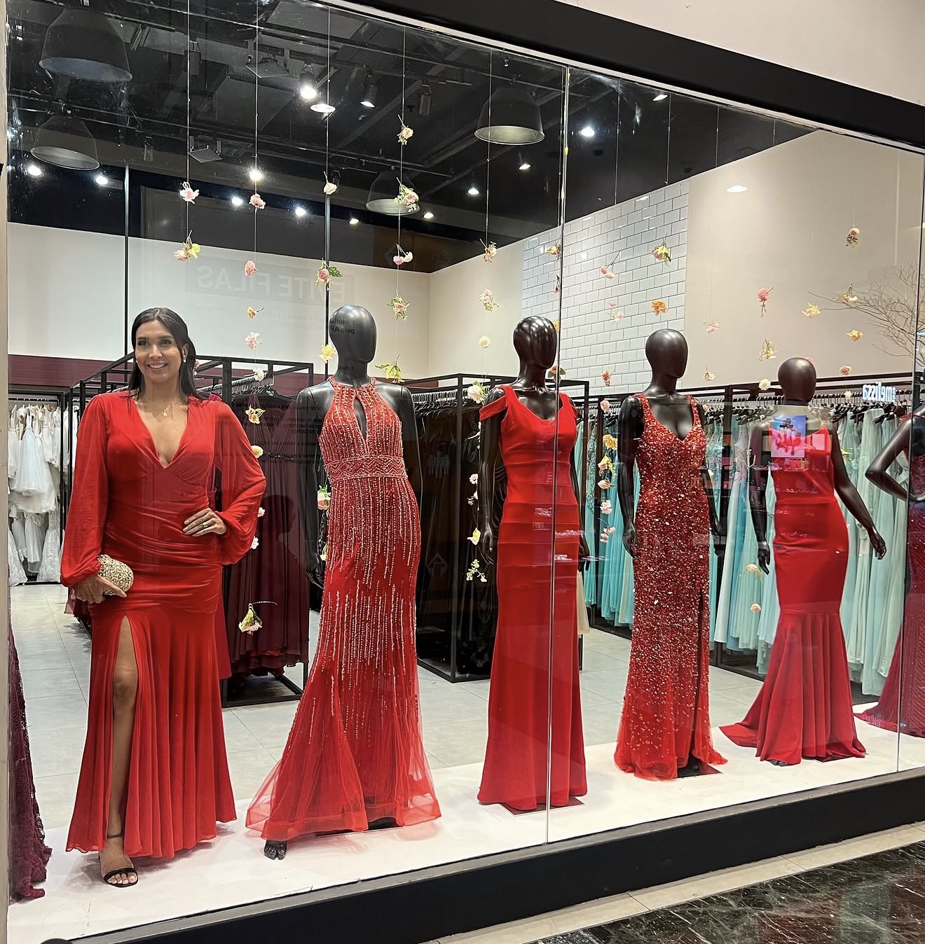 Woman in red dress with pleated skirt and slit, standing in front of mannequins dressed in red evening gowns inside a store window.