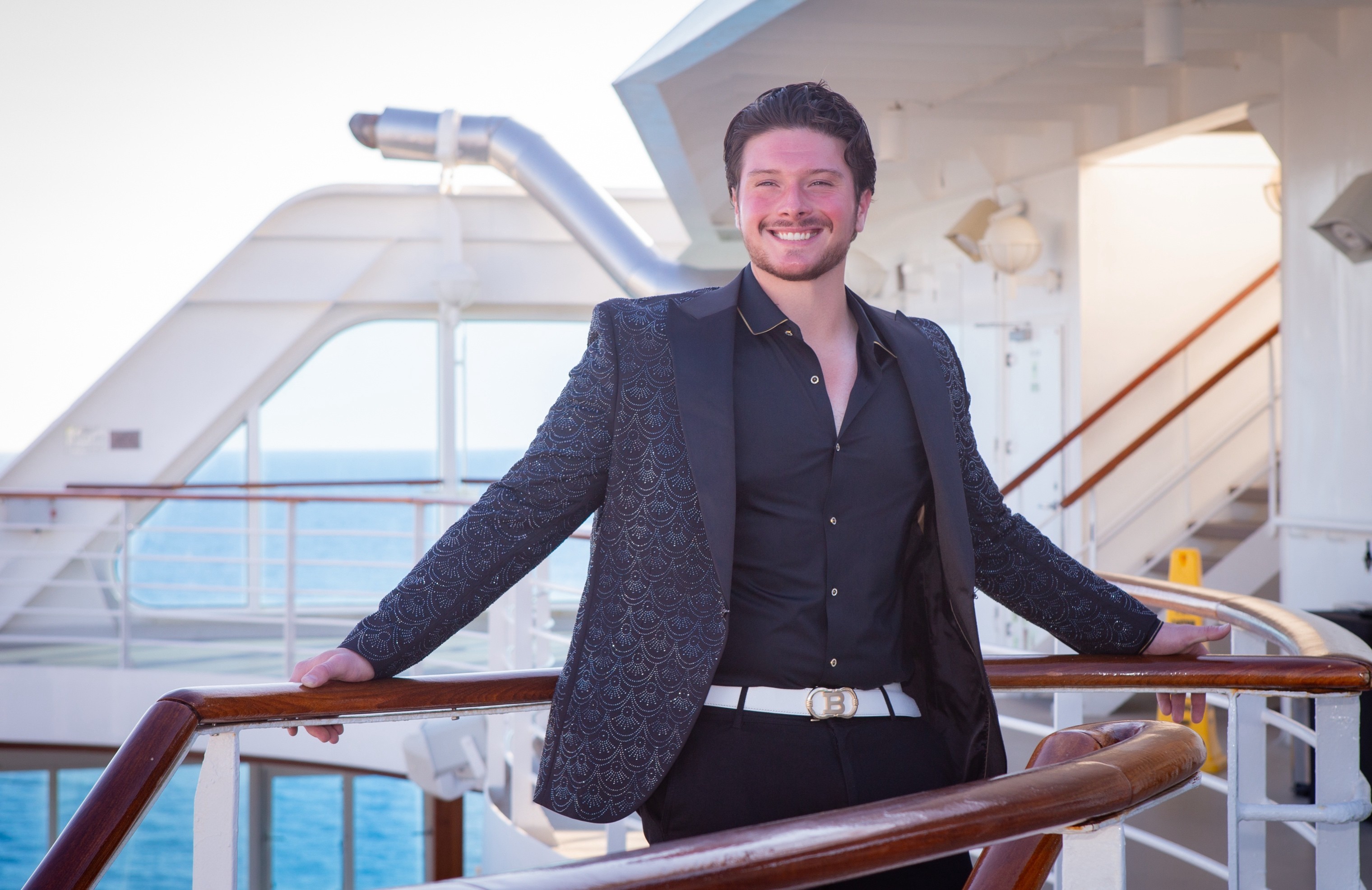 Man with dark hair and beard smiling, wearing a dark blazer and shirt, standing outdoors on a ship deck with railings and stairs.