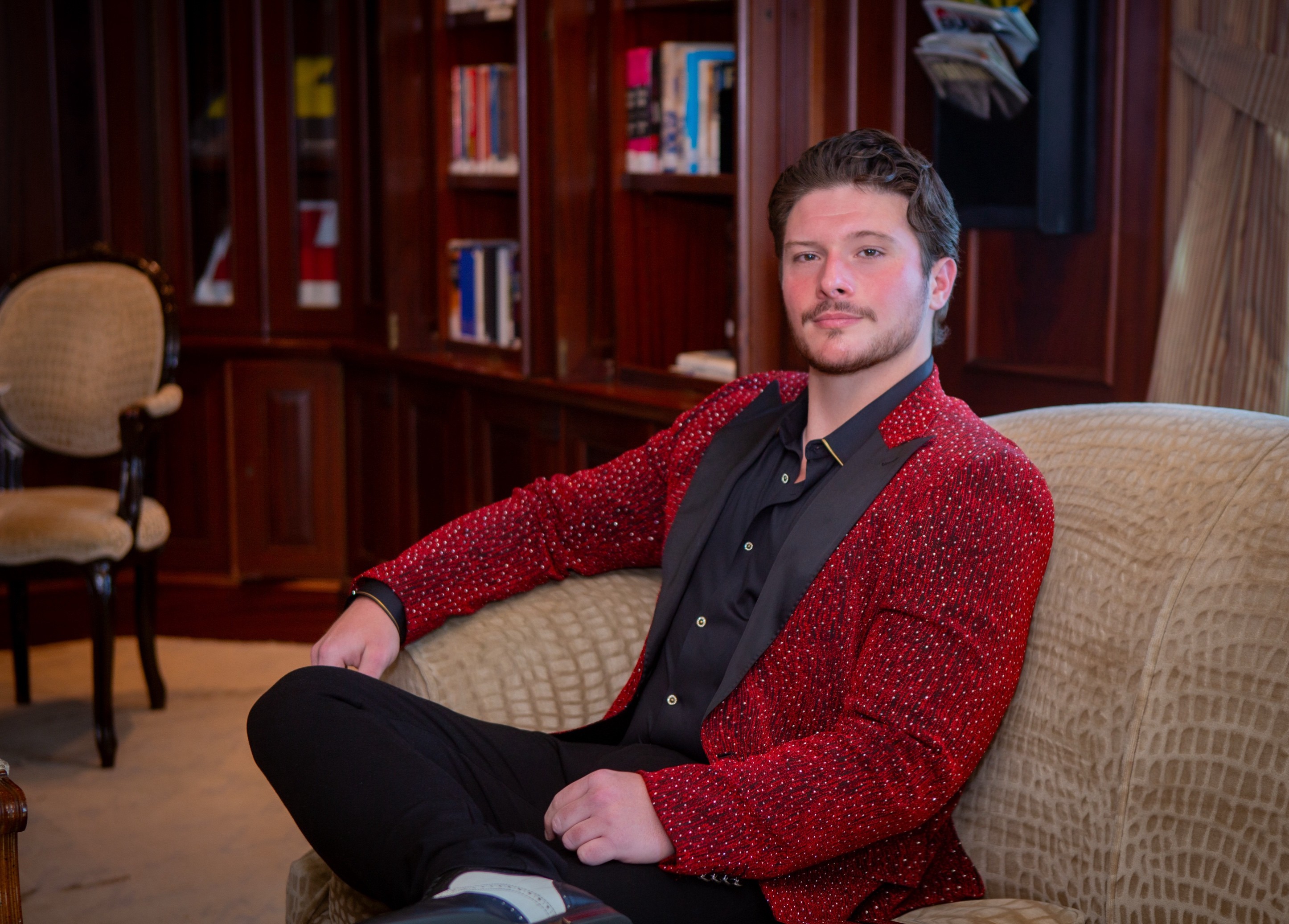 Young man with dark hair and beard sitting on a beige sofa in a room with wooden bookshelves, wearing a red jacket and black shirt.