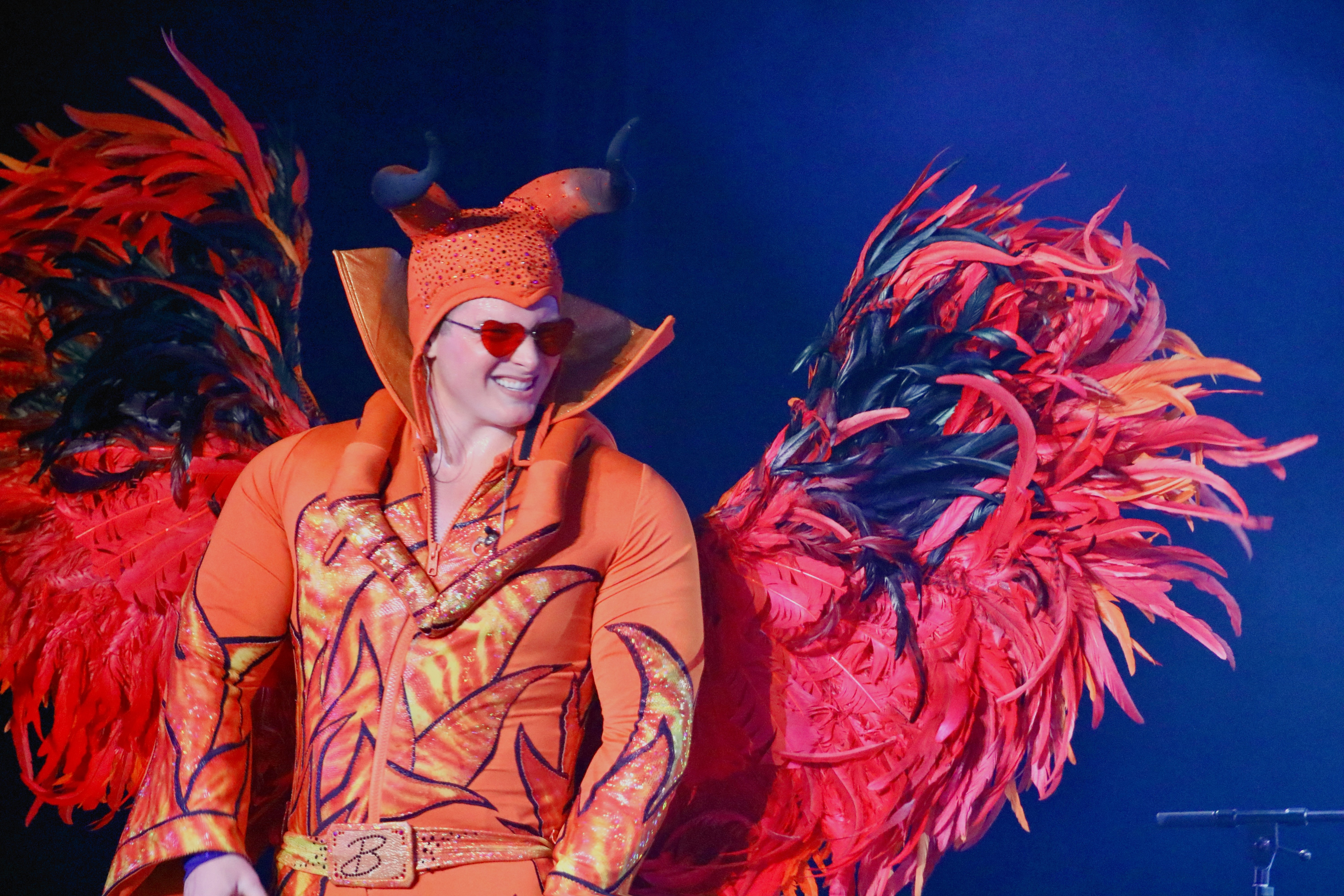 Person in colorful costume with large red and black feathered wings, wearing a hat with horns and sunglasses, smiling.