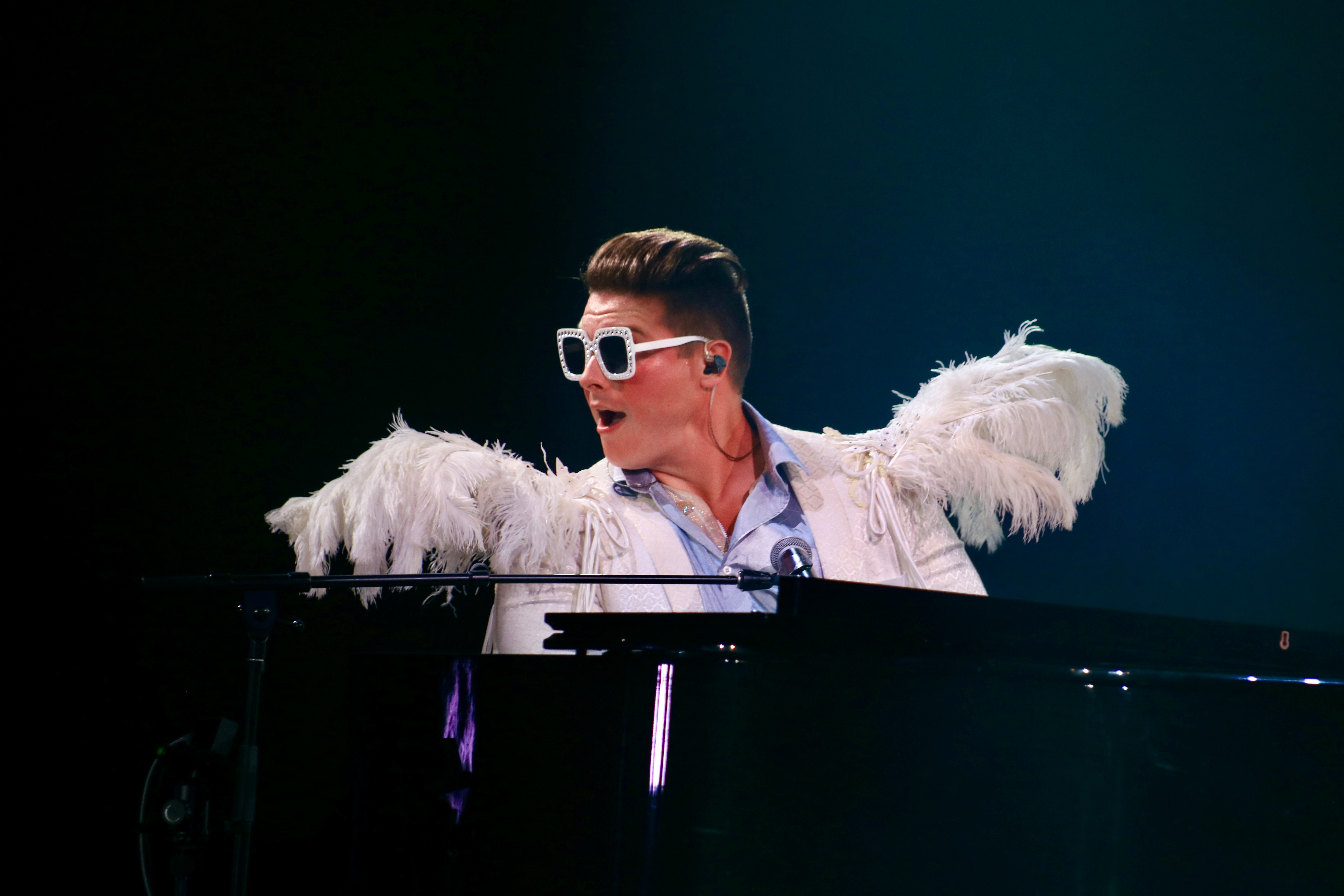 Person wearing sunglasses and a feathered jacket playing a piano on stage, dark background.