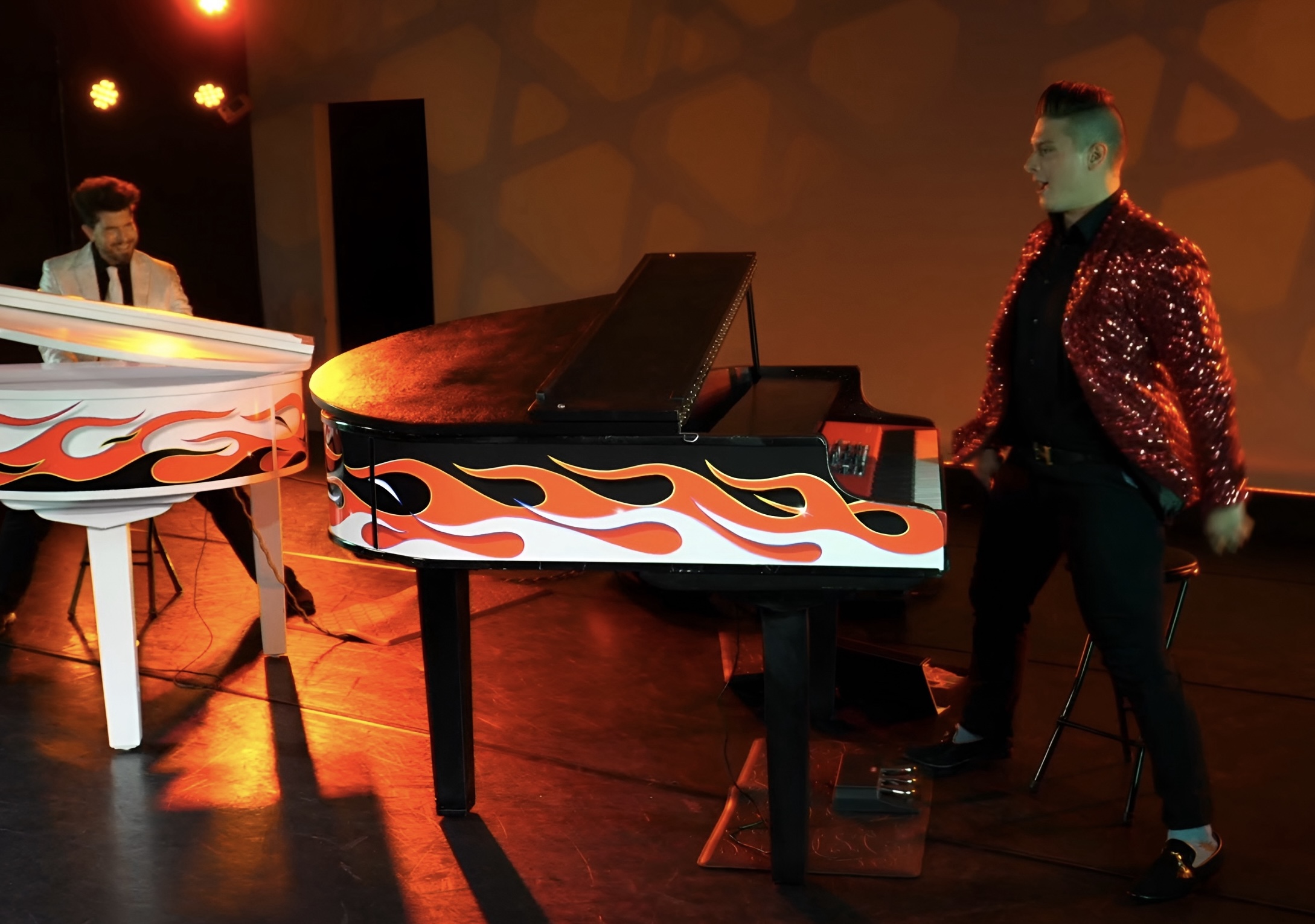 Two performers on stage with a decorated piano, one in a white jacket with flames, the other in a red sequined jacket.