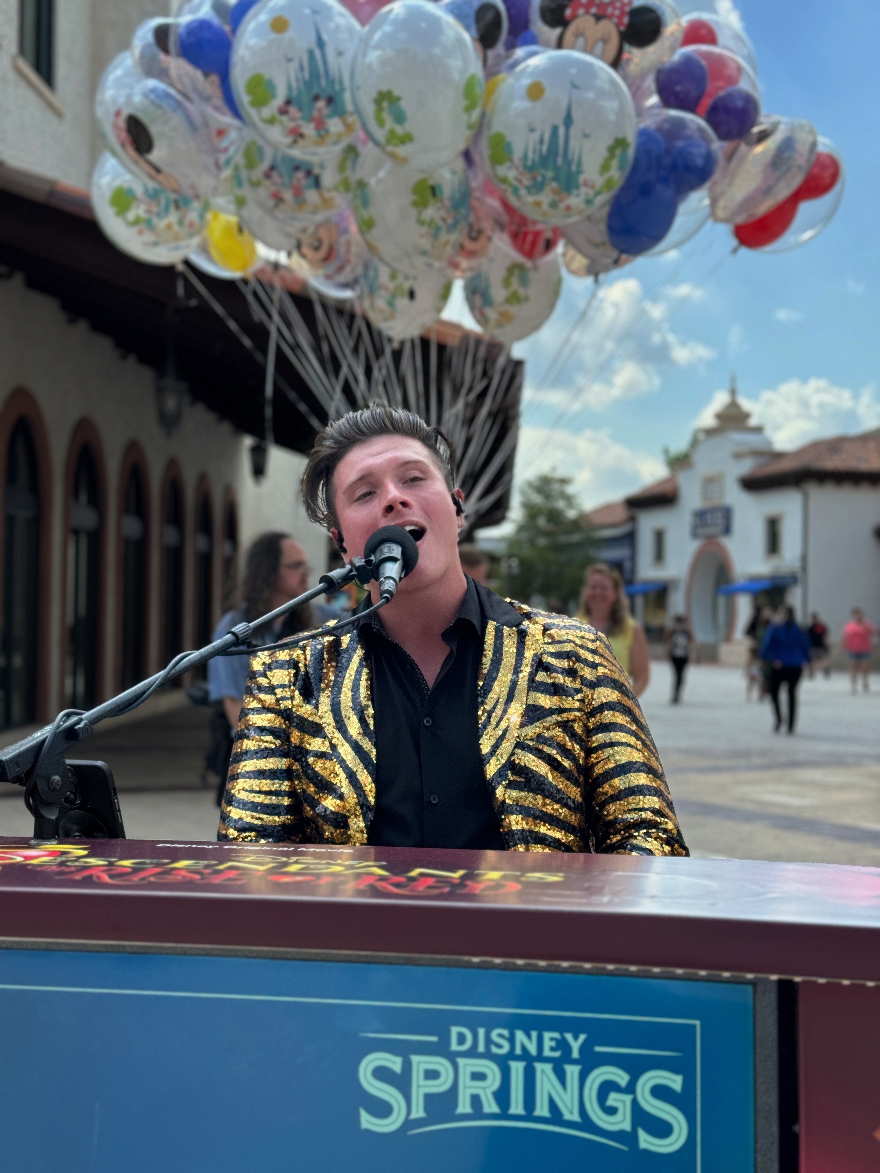 Person singing into a microphone with balloons above, outdoors in a plaza with buildings and people in background.