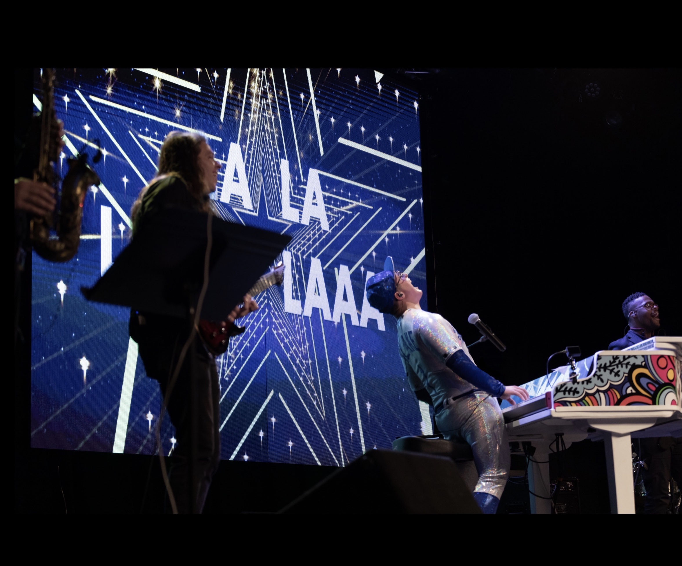 Musicians perform on stage with a large screen displaying text and star graphics in the background.
