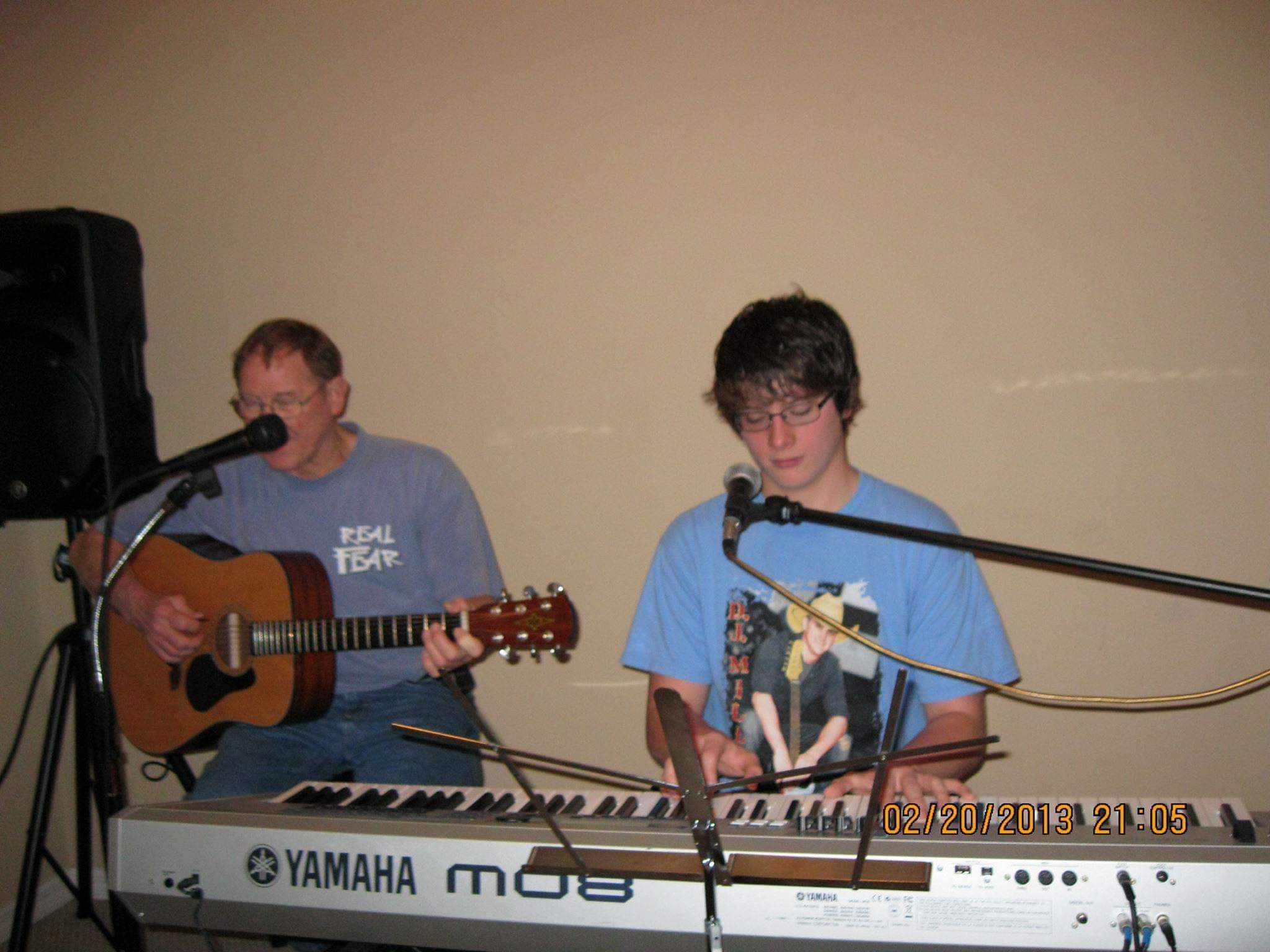 Two people perform music; one plays guitar, the other plays keyboard, both with microphones, against a plain background.