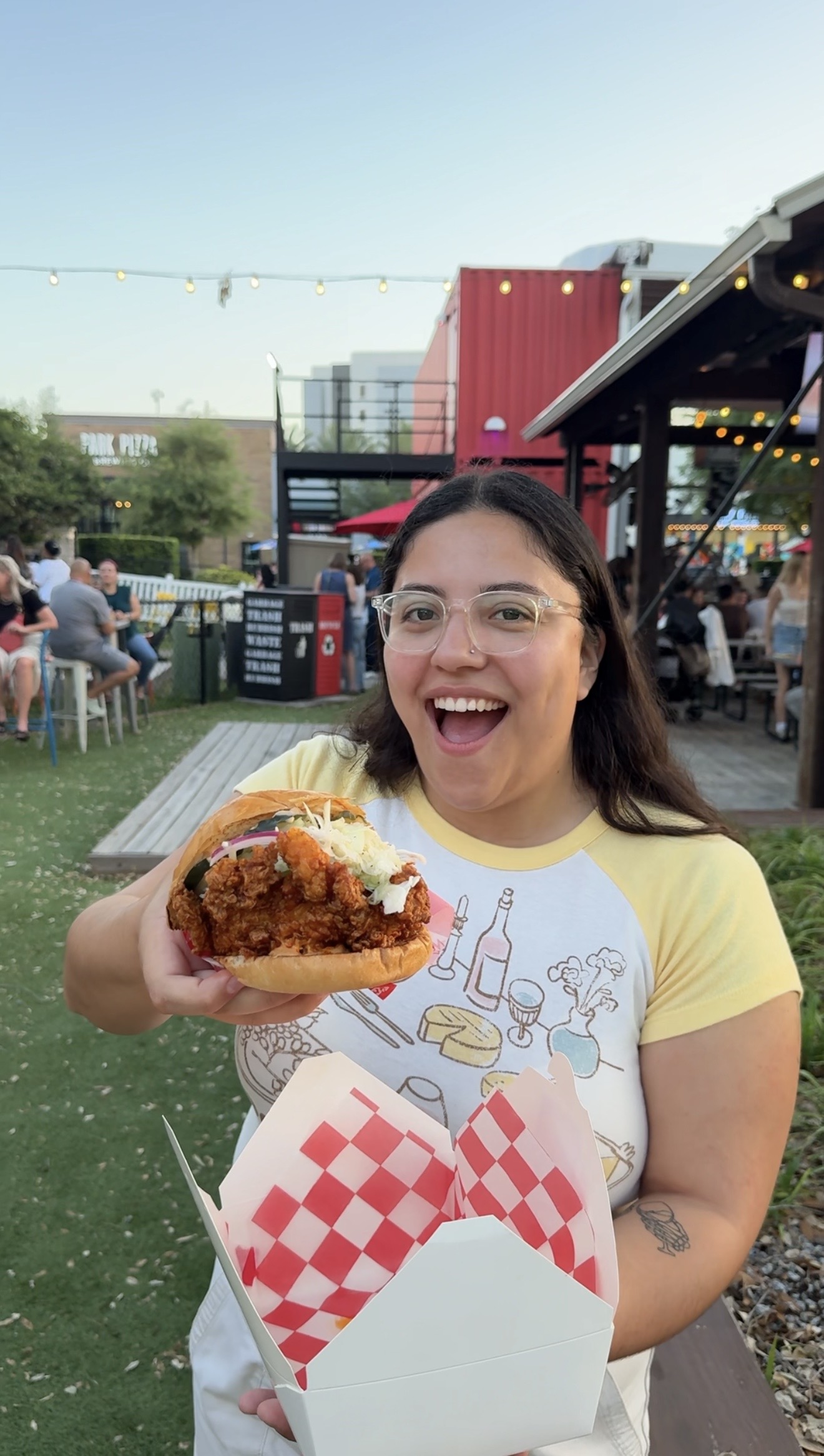 Woman smiling and holding a large taco with toppings outdoors, with people and structures in the background.