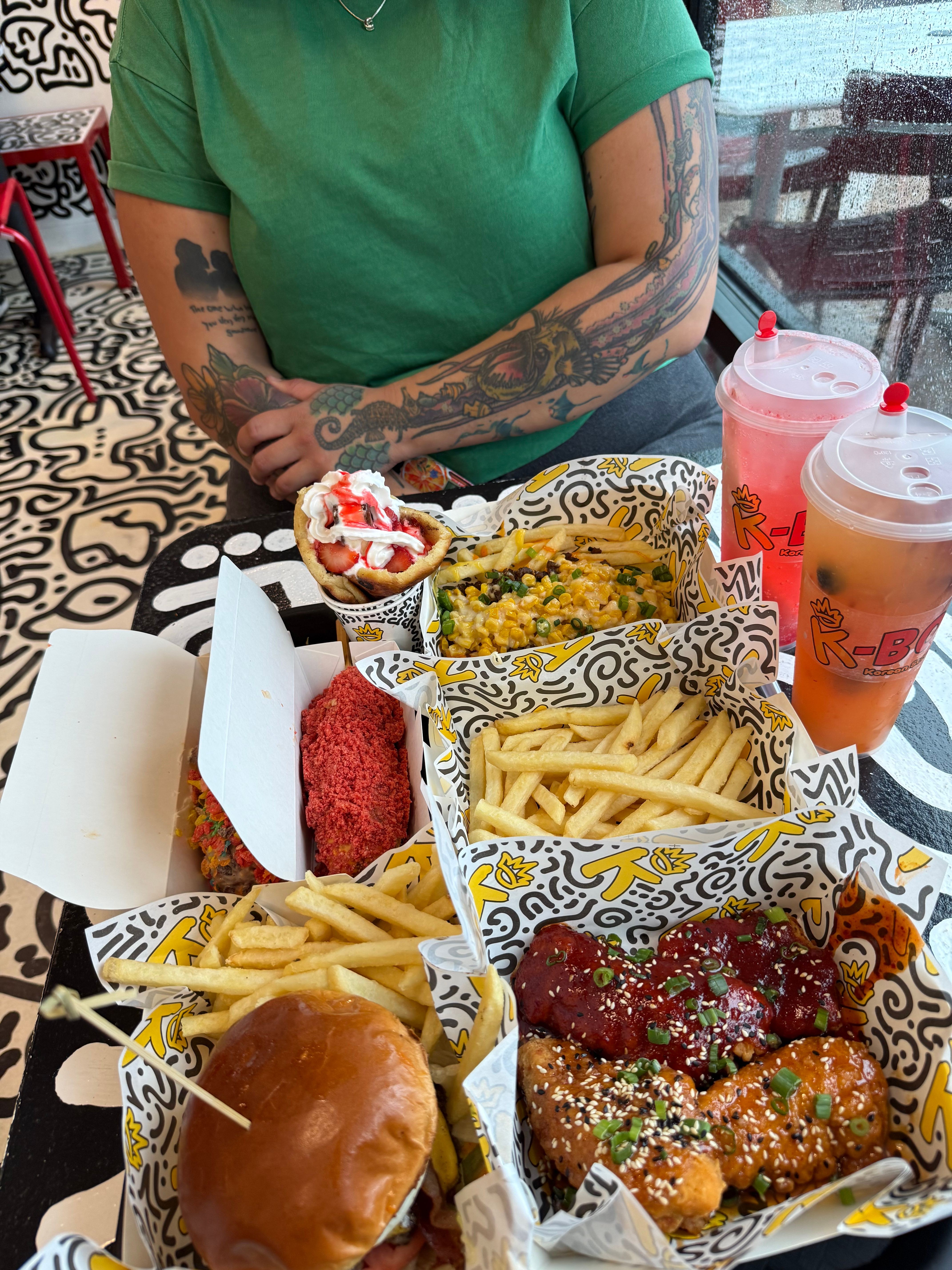 Person with tattoos holding a cupcake, surrounded by fries, chicken, and drinks on a table.