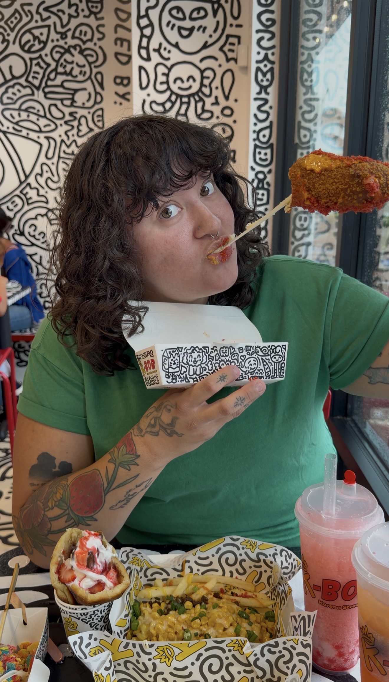 Person with curly hair in green shirt eating food with a skewer, sitting at a table with food and drinks, background with patterned wall.