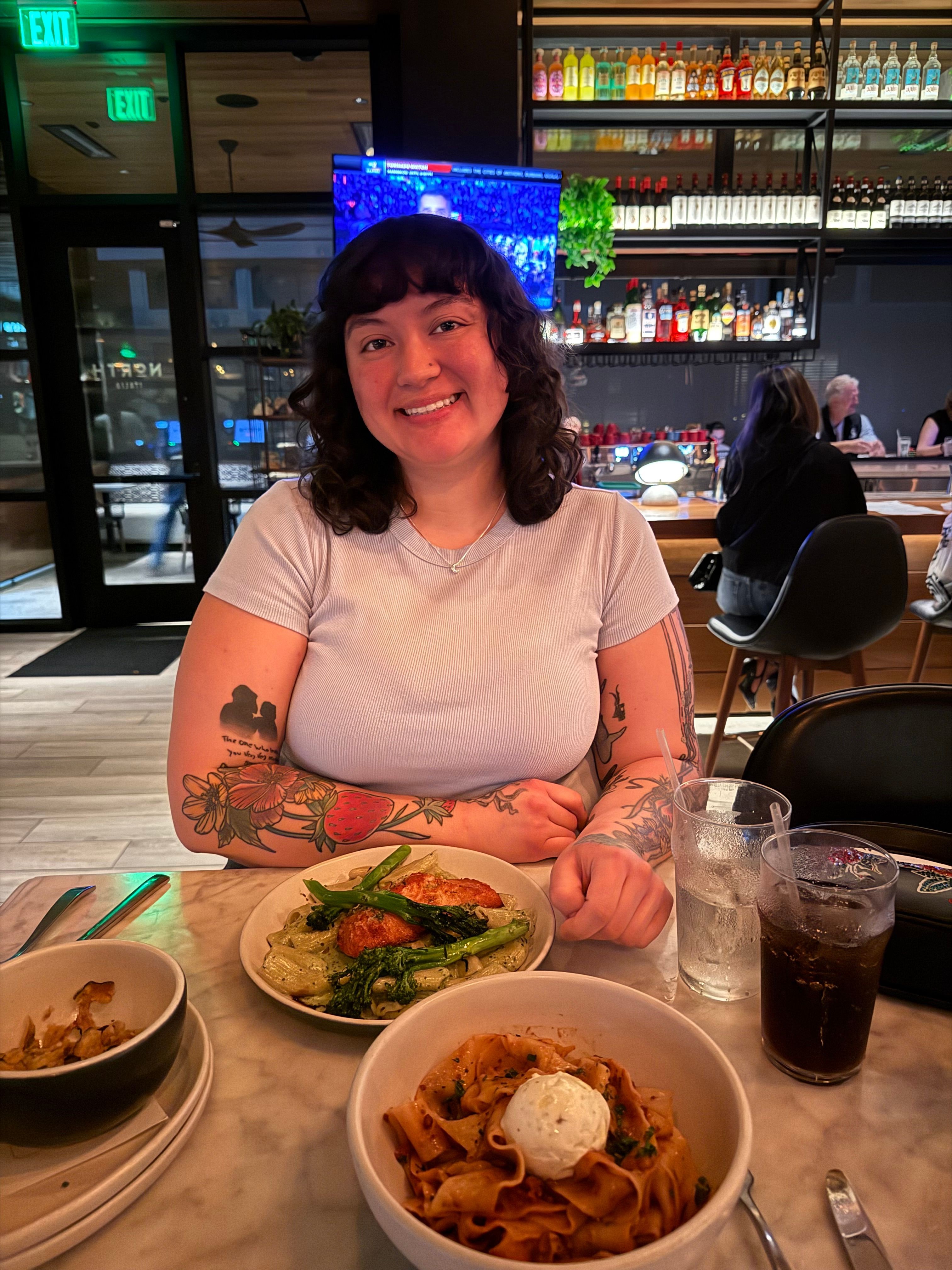 Woman with dark curly hair and tattoos on her arm, smiling, sitting at a table with pasta and salad, in a restaurant.