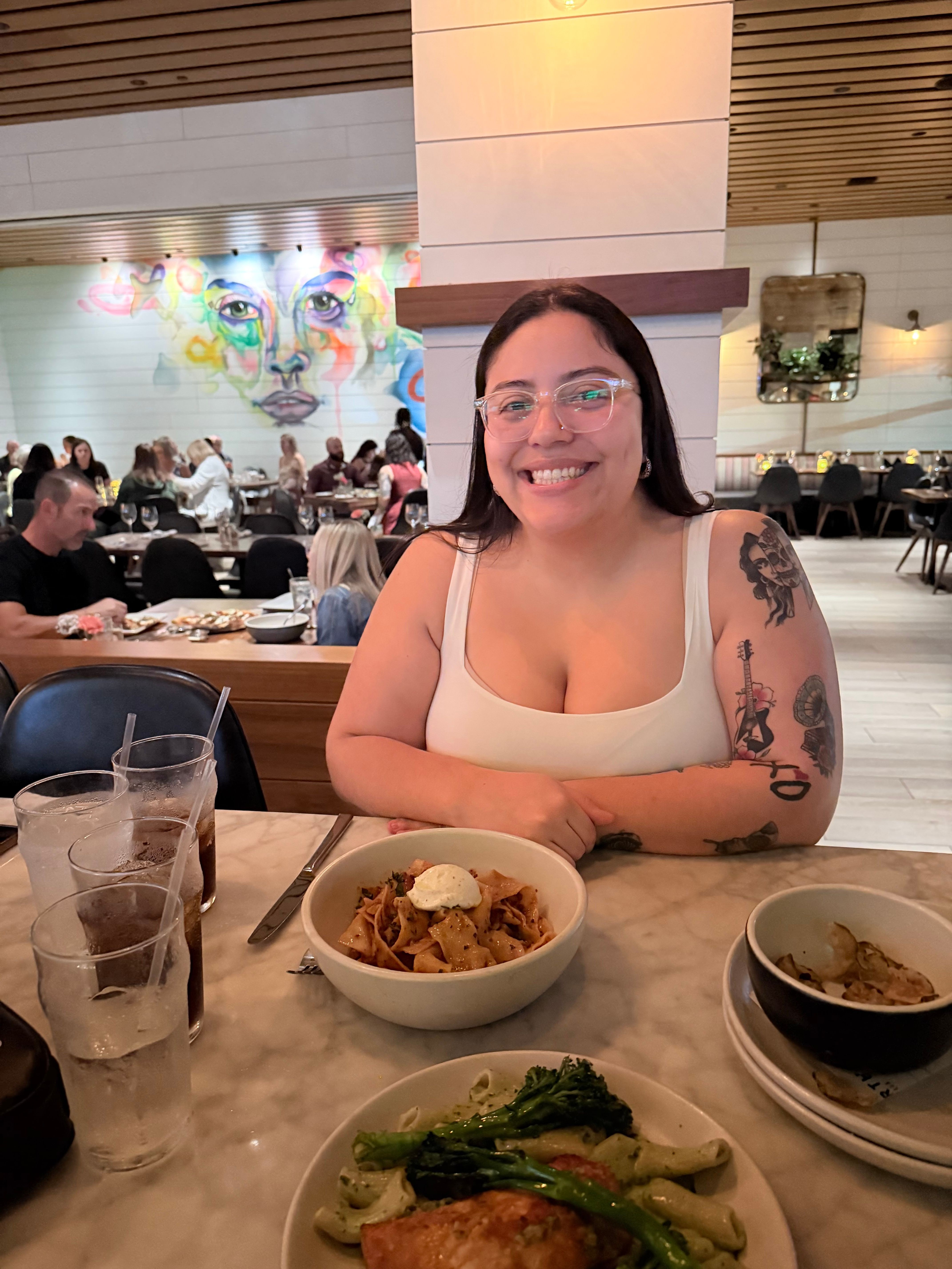 Woman with glasses and tattoos smiling at a restaurant table with bowls of food and drinks.