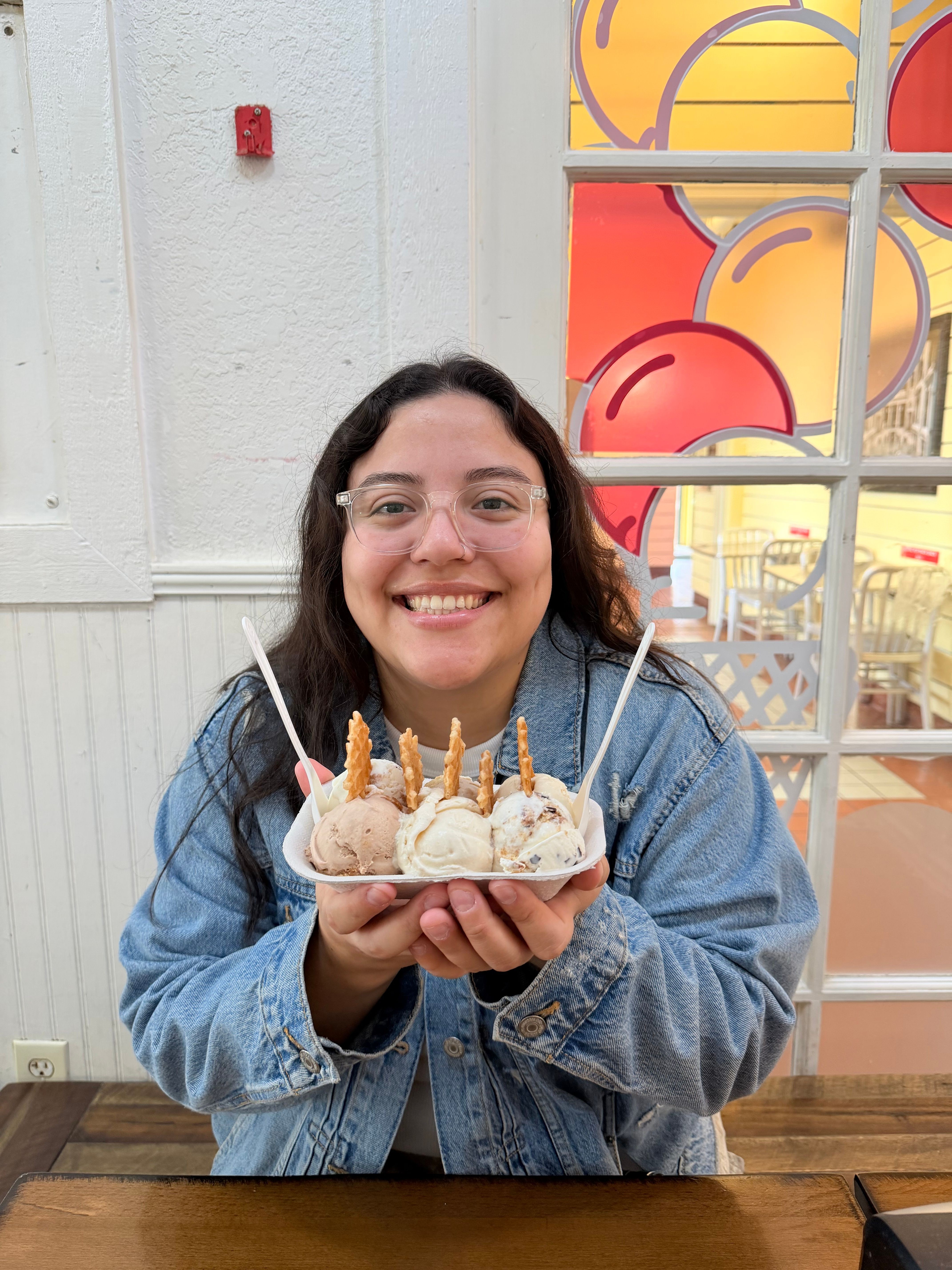 Woman smiling and holding a birthday cake with lit candles, sitting at a table indoors.