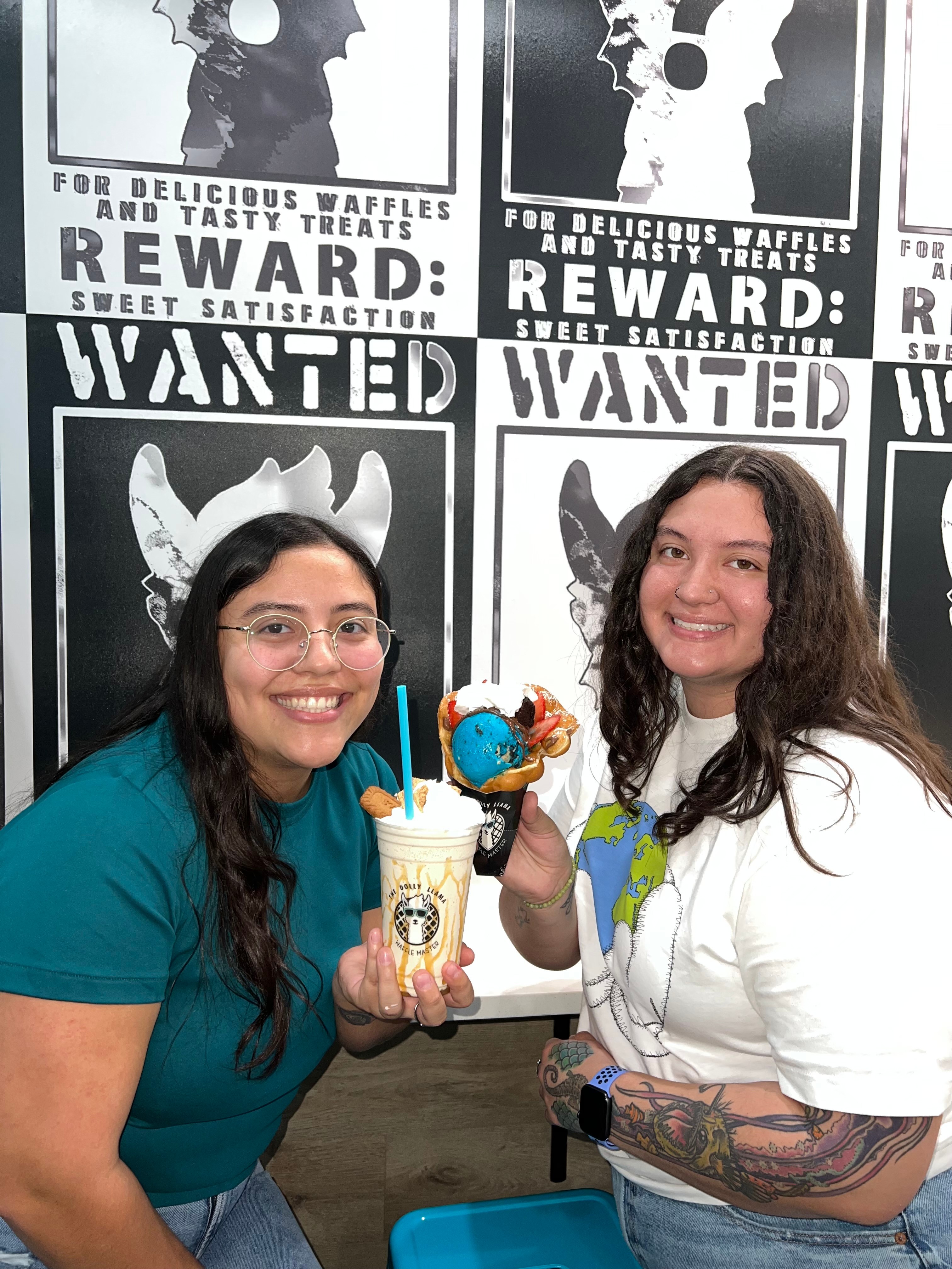 Two women smiling and holding a small plush toy and a drink, standing in front of a black and white wanted poster wall.