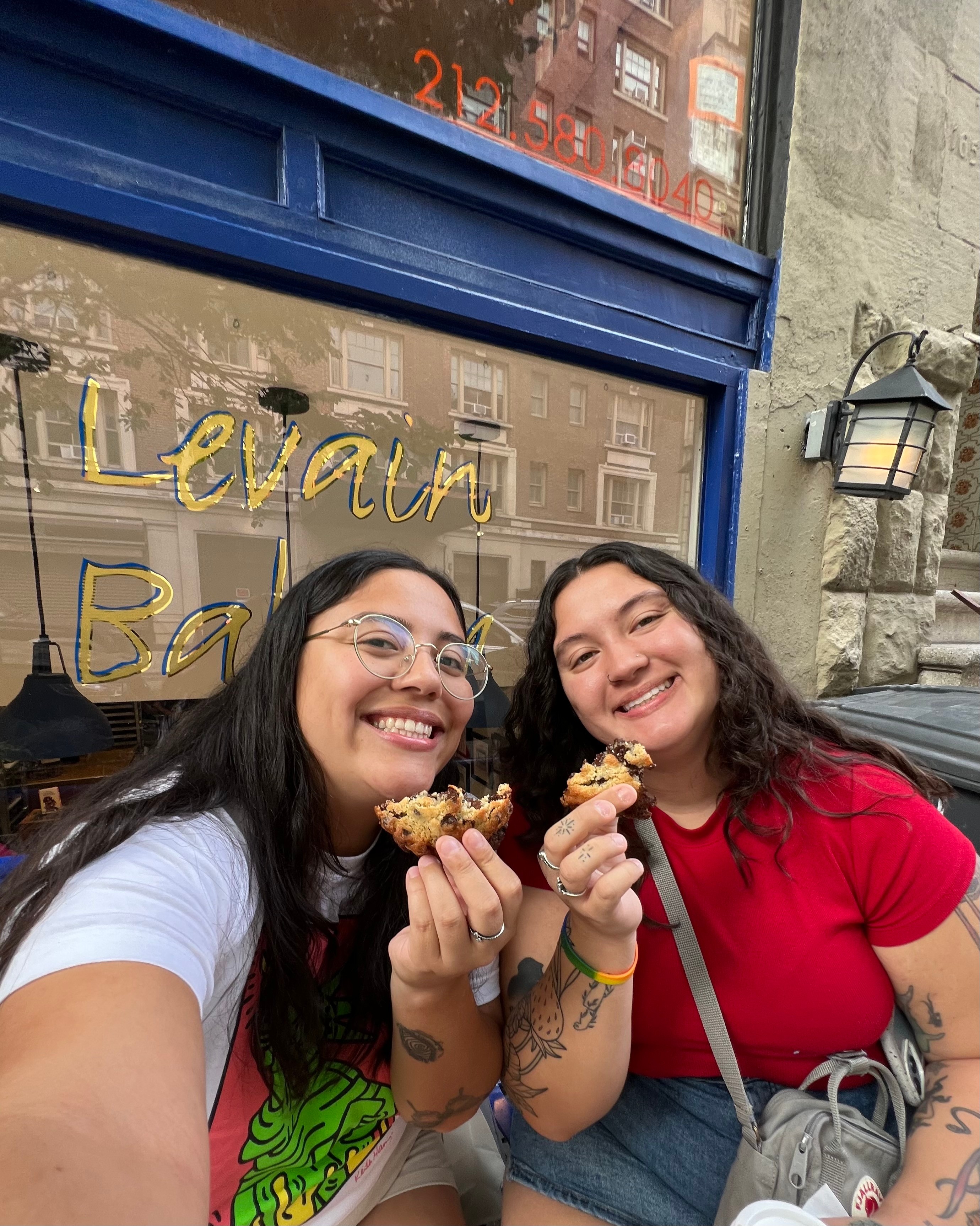 Two women smiling and holding ice cream cones outside a shop with a blue window frame and a stone wall.