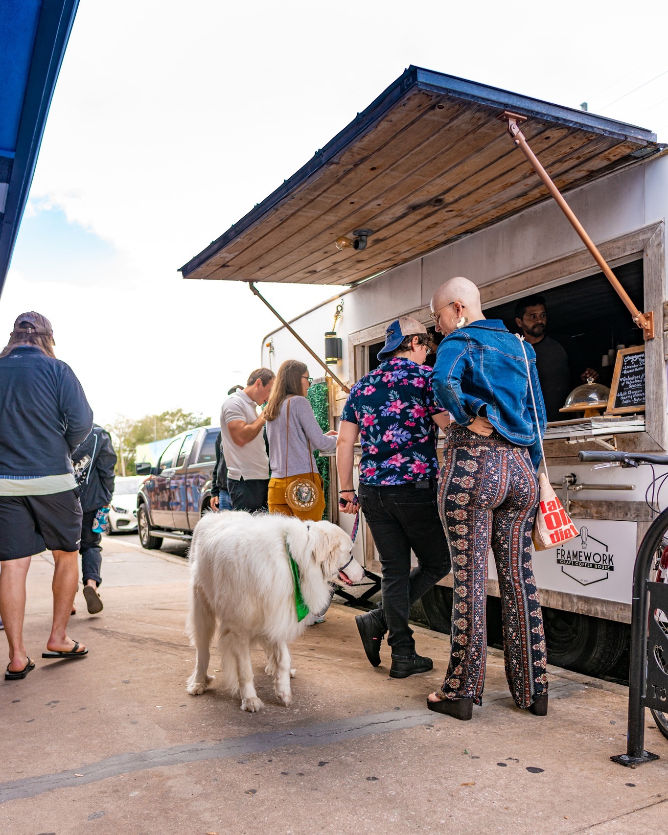 People line up at a food truck with a dog on a leash, under a wooden awning, in an outdoor setting.