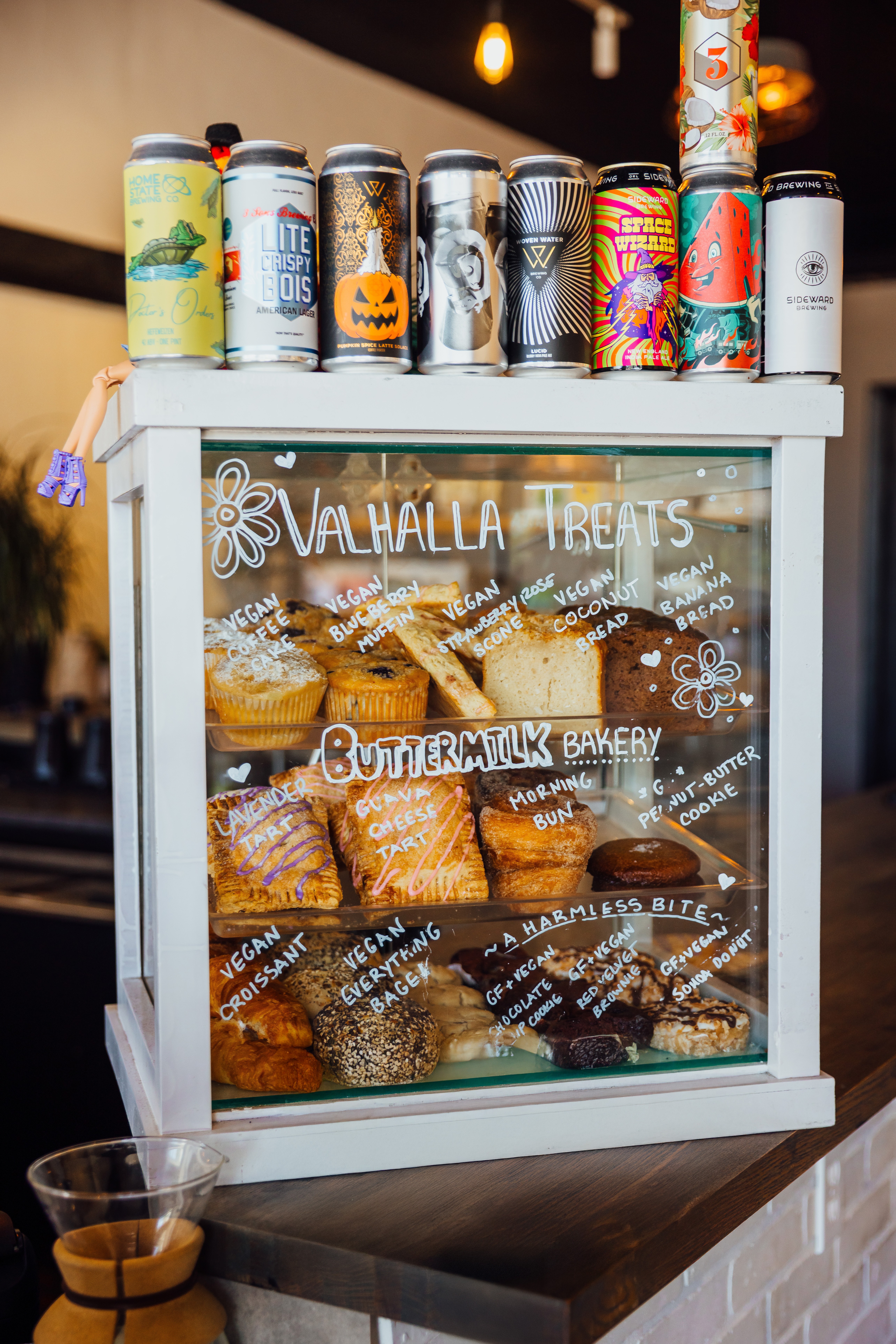 Glass display case with baked goods and handwritten signs, topped with colorful cans and bottles, in a bakery setting.