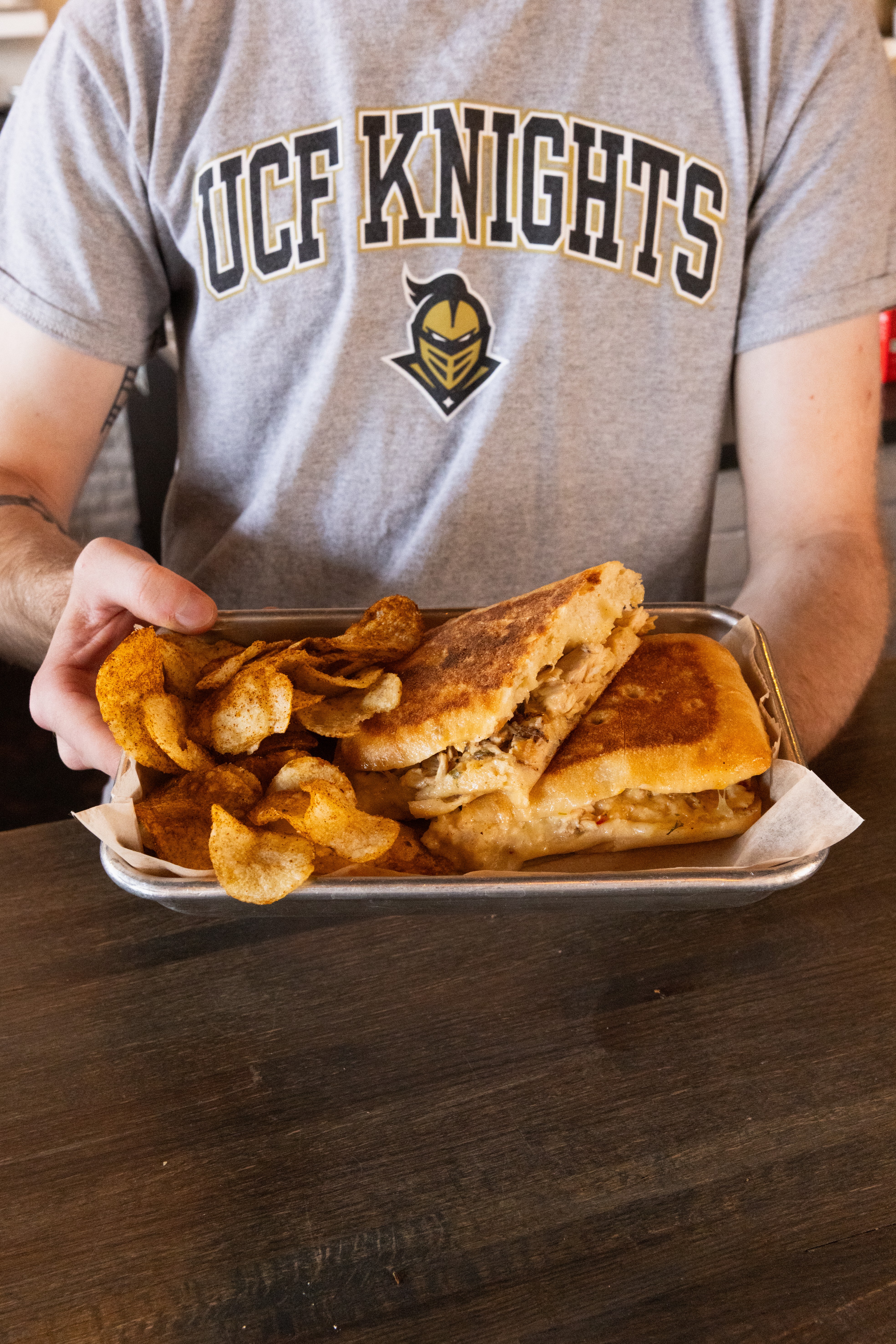 Person wearing a gray UCF Knights shirt holding a tray of fried chicken, biscuits, and gravy, sitting at a dark table.