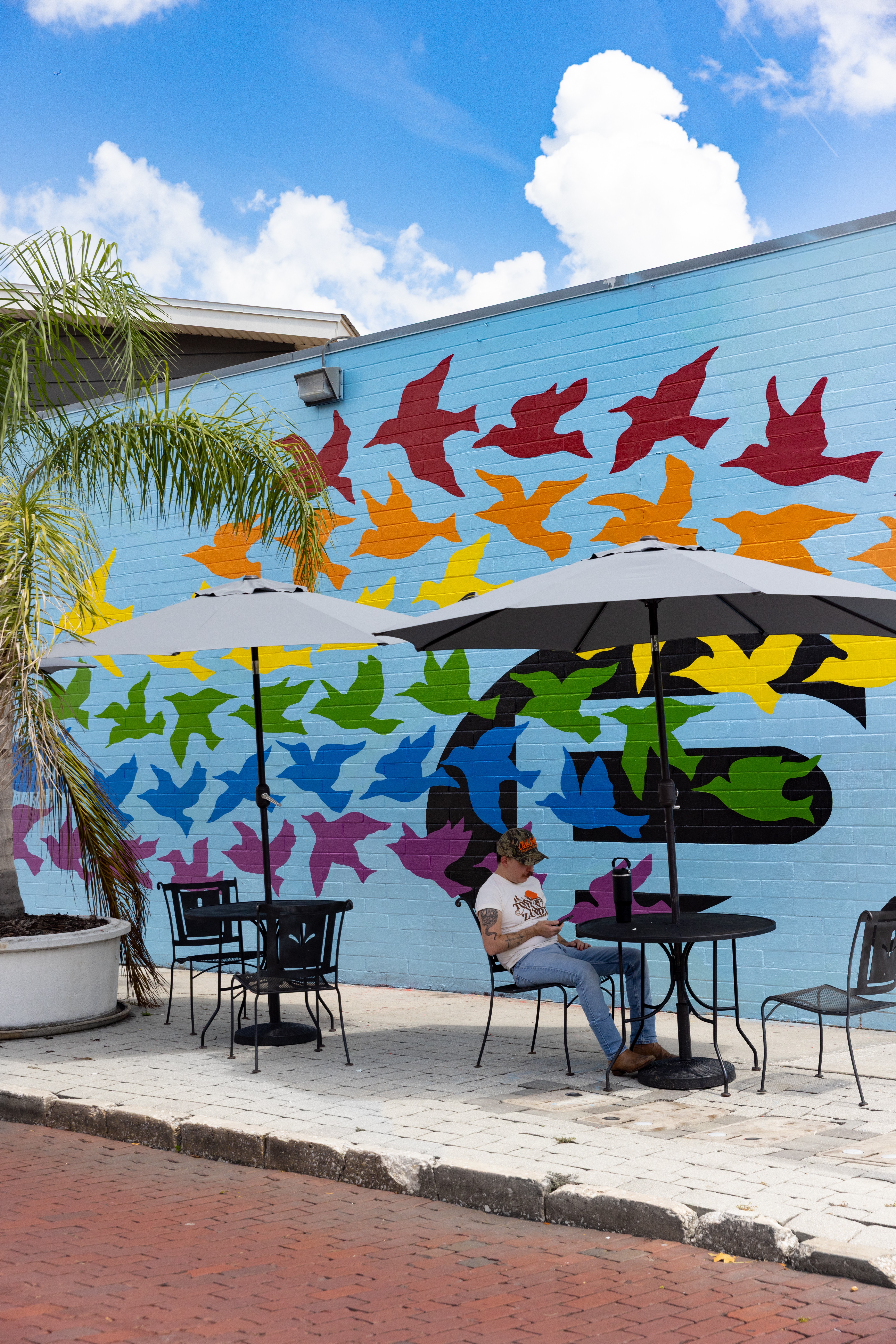 Outdoor seating area with tables, chairs, umbrellas, and a colorful bird mural on the wall, under a blue sky with clouds.