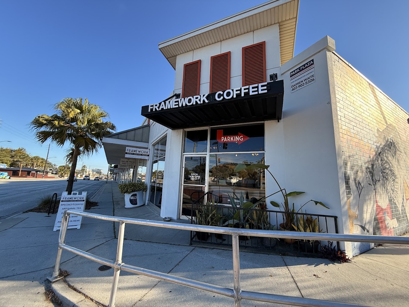 Corner of a coffee shop with a sign reading 'FRAMEWORK COFFEE', a palm tree, and a clear blue sky.