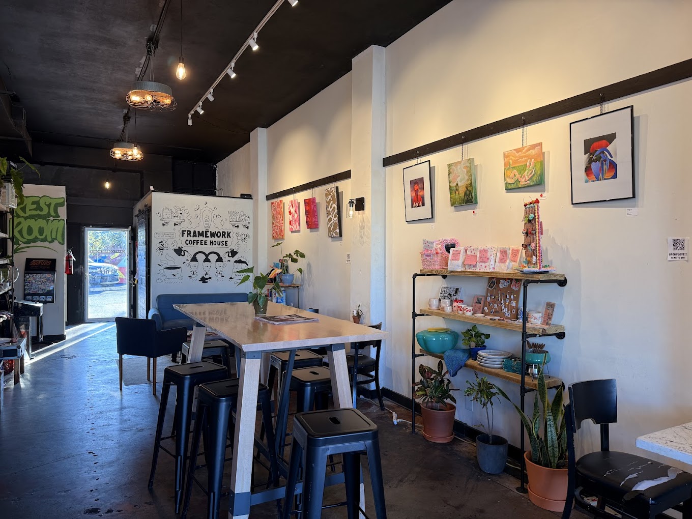Interior of a cafe with artwork on white walls, a wooden table, chairs, and shelves with plants and decorations.