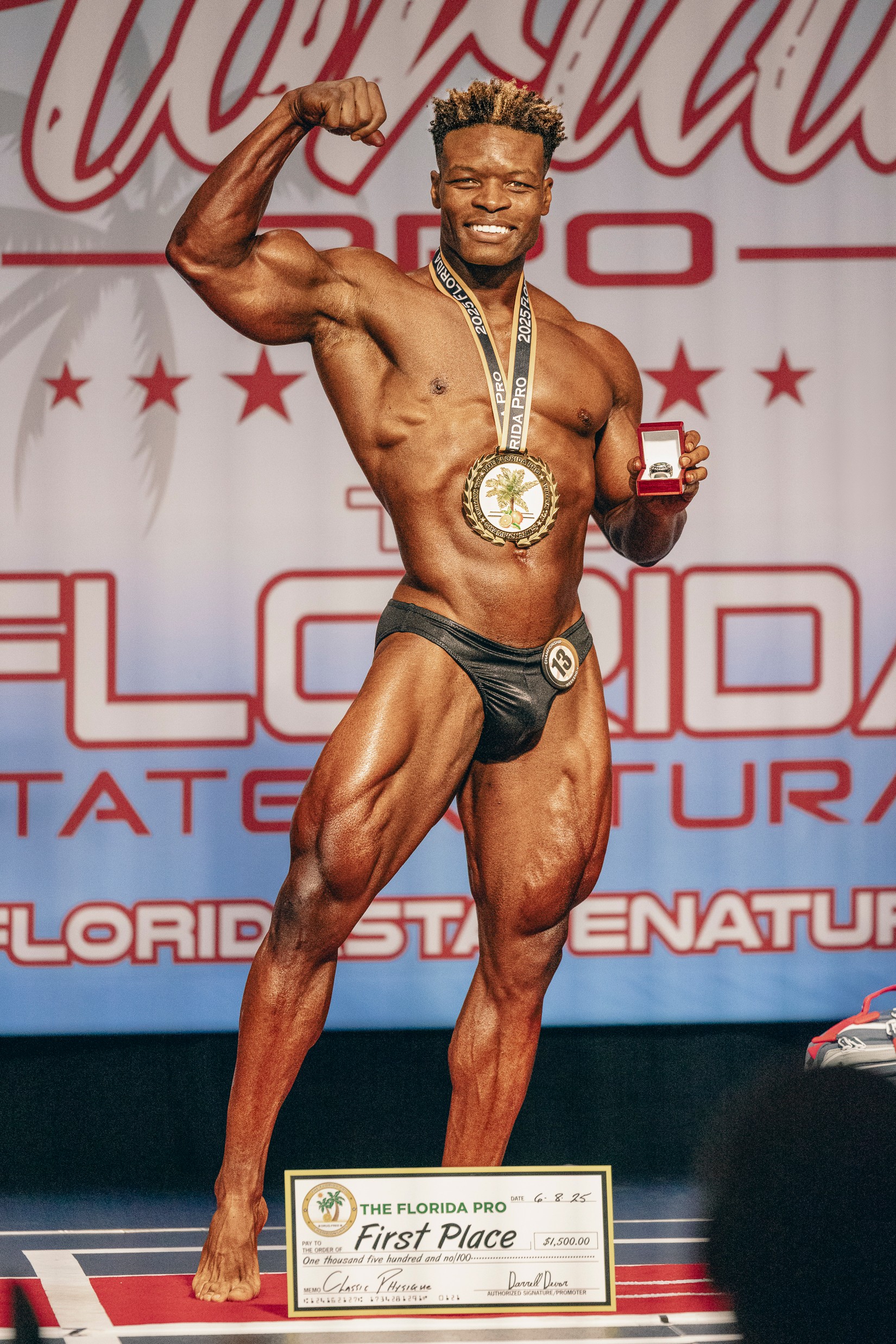 Male bodybuilder on stage flexing, holding a medal and a small trophy, smiling, with a backdrop and a first-place plaque.