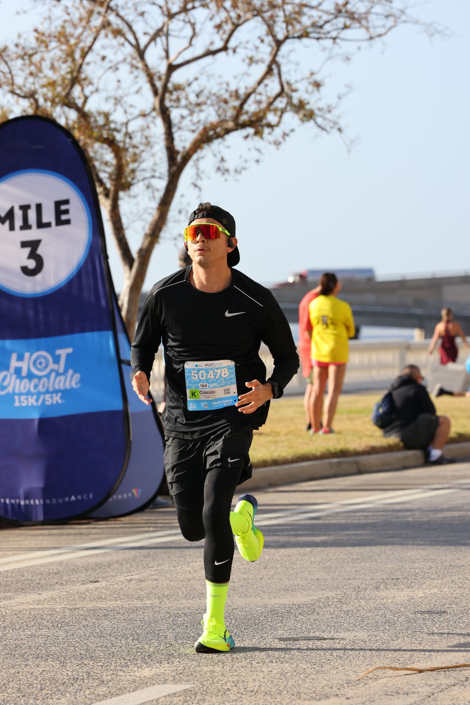 Runner in black outfit and neon shoes holding a water bottle, with a blue sign and trees in the background.