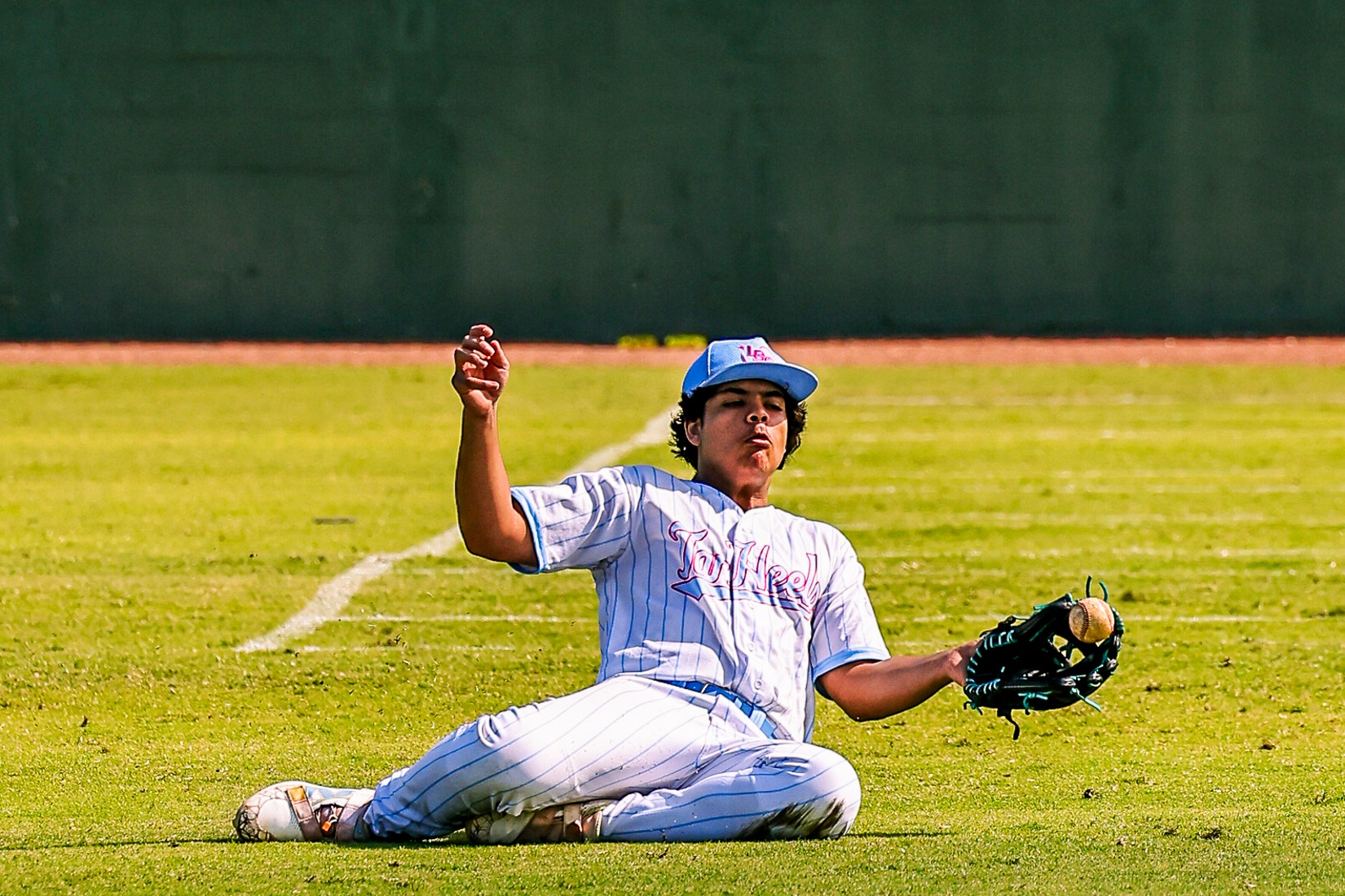 Baseball player sitting on grass field, holding glove and ball, with a green wall in background.