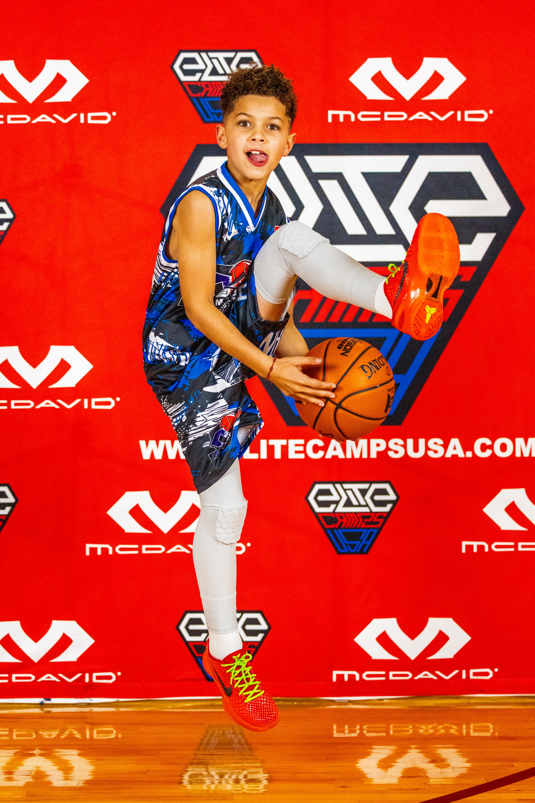 Child in basketball uniform holding a basketball, smiling, against a red background with logos.