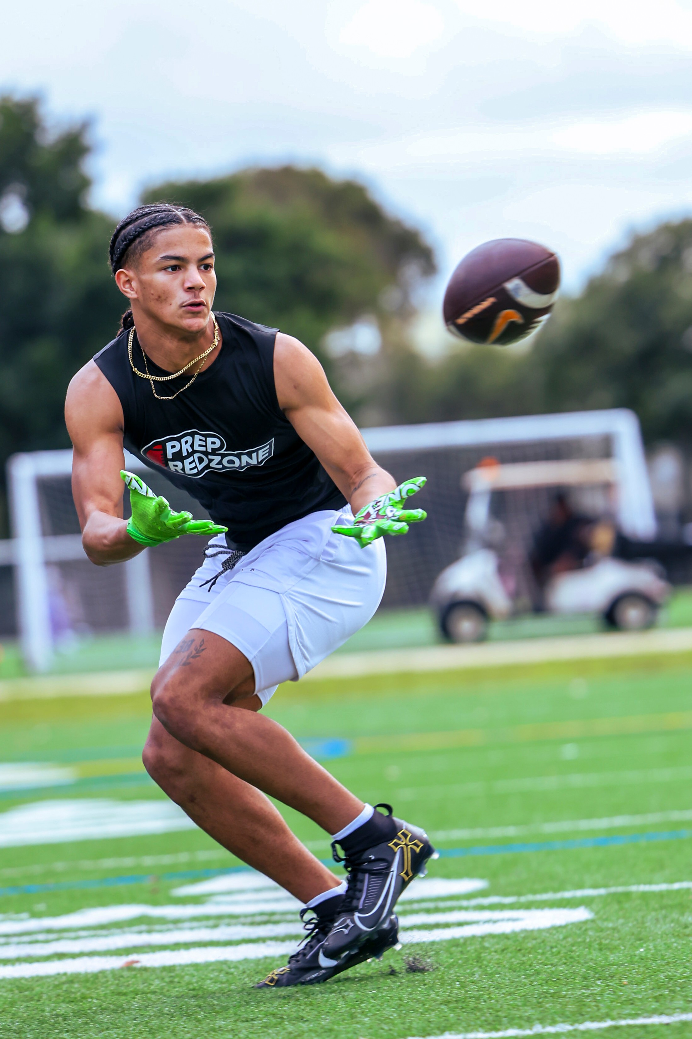 Young athlete in black shirt and white shorts catches football on field with trees and vehicles in background.