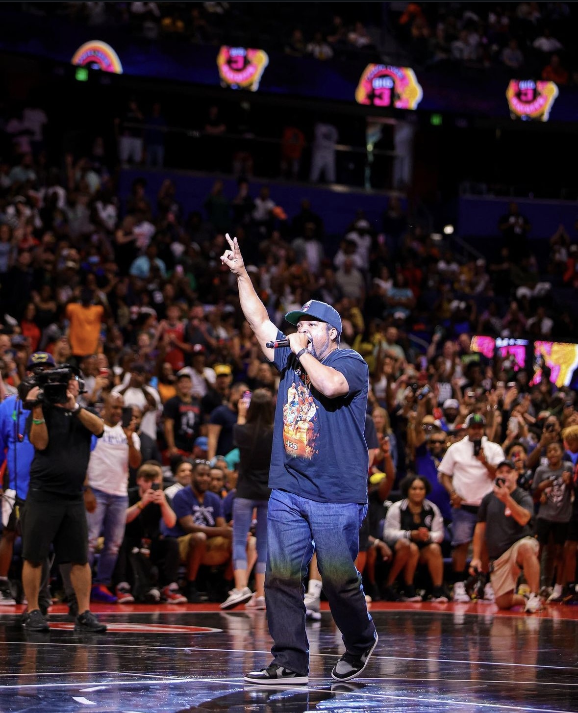 Person with a cap and T-shirt speaking into a microphone on a basketball court, crowd in the background.