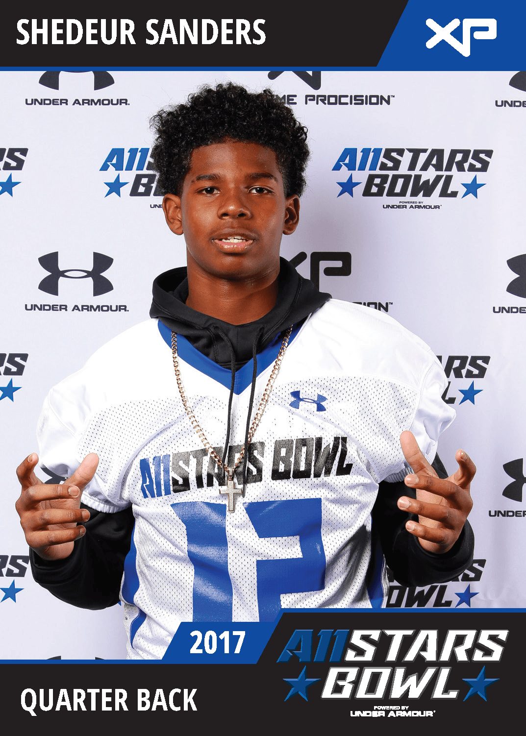 Young football player in uniform holding two white helmets, standing in front of a backdrop with logos, wearing a medal.