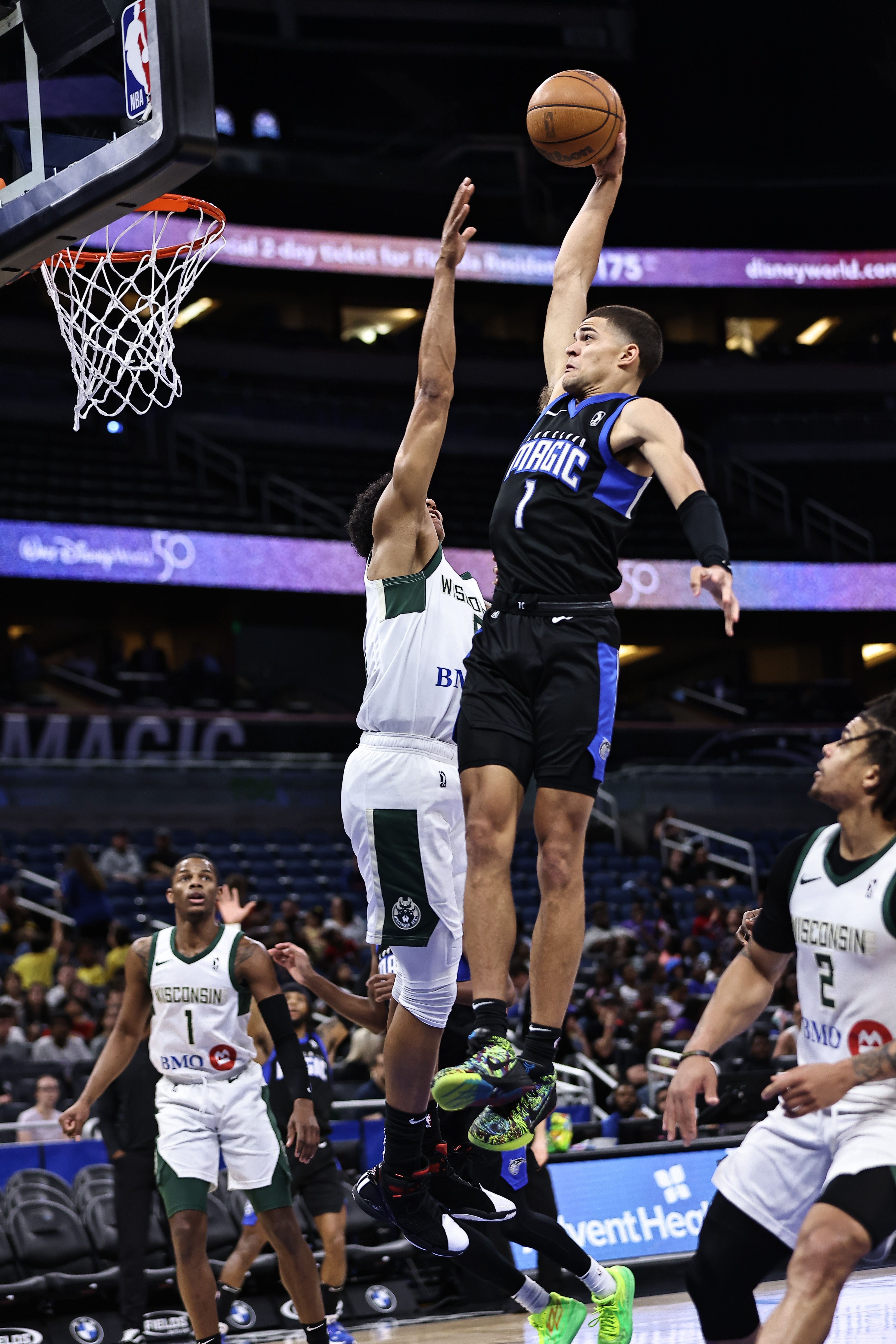 Basketball player in black jumps to shoot, others watch, in an indoor arena with spectators and digital screens.