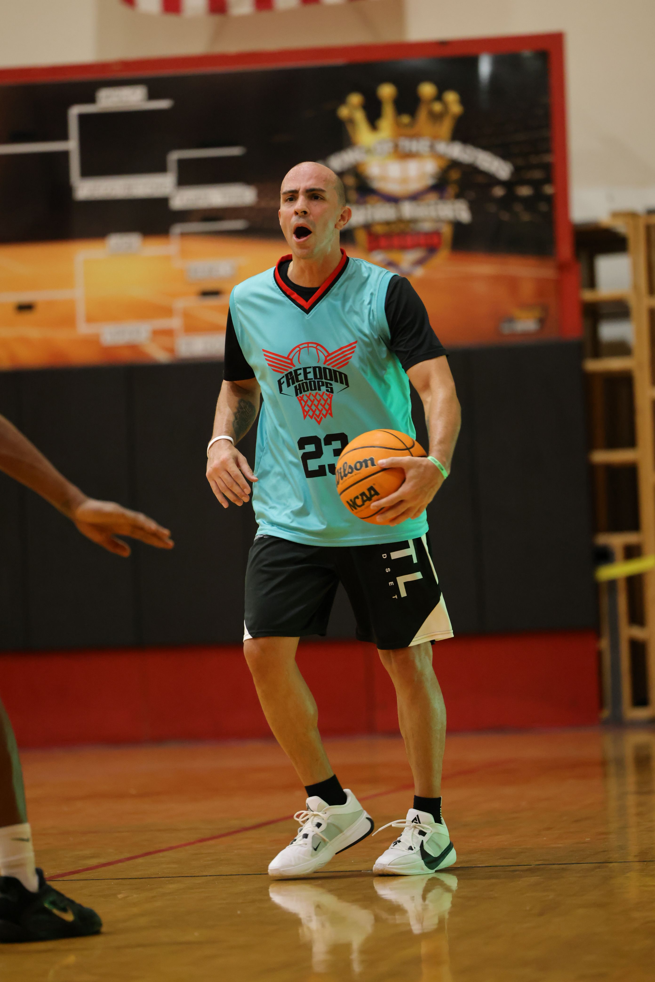 Person holding a basketball on an indoor court, wearing a light blue jersey and black shorts, with a red and black sleeve on one arm.