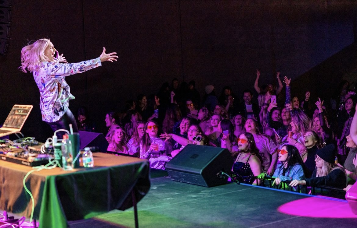 Performer on stage with arms outstretched facing an audience in a dark room, colorful lighting, and a table with equipment.