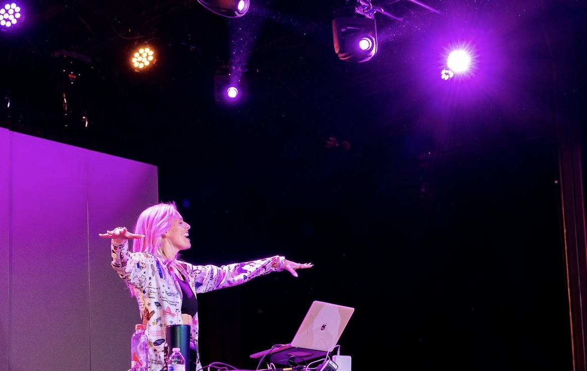 Performer with pink hair pointing on stage under purple and pink lights, with a laptop and equipment nearby.