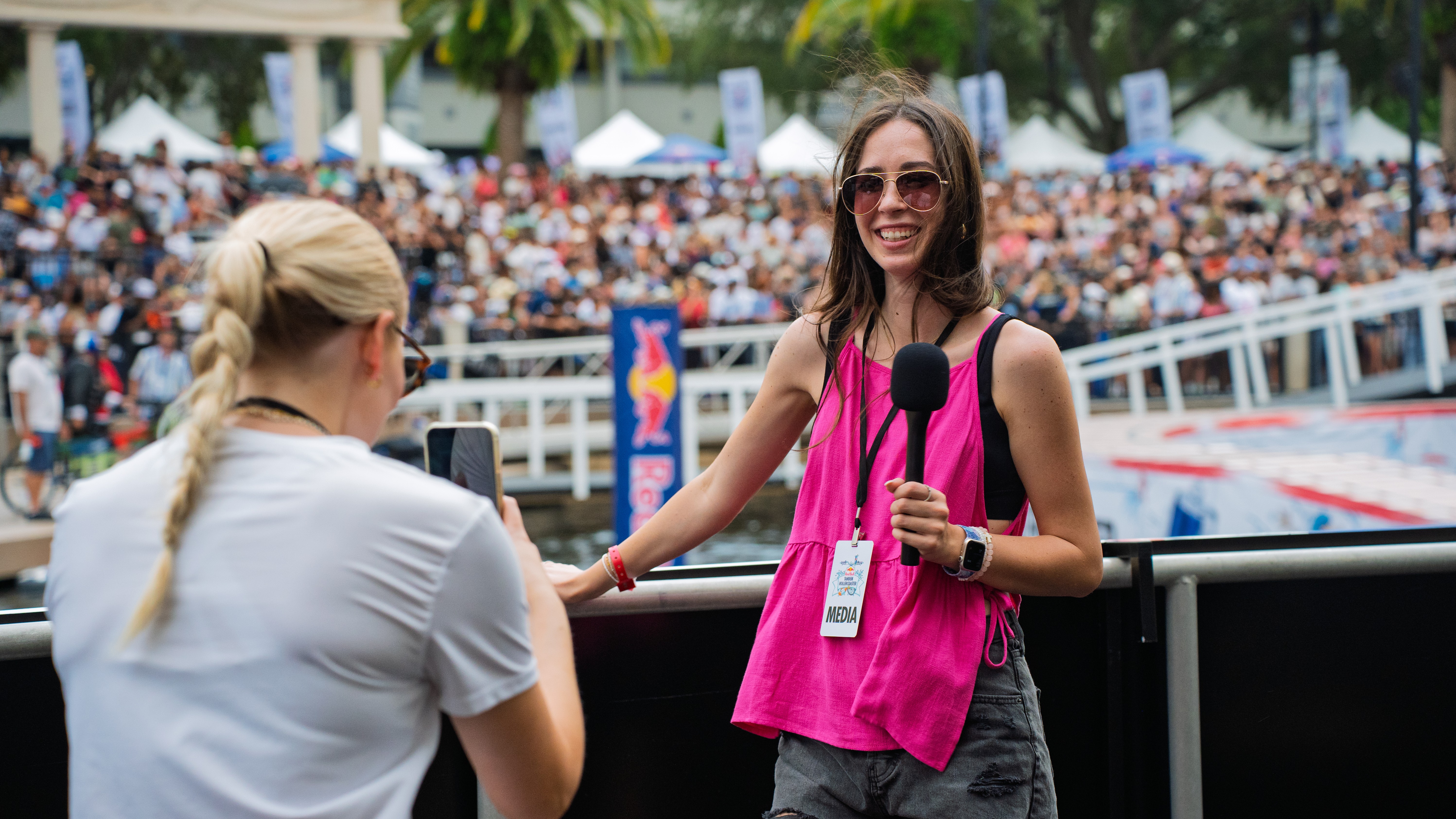 Woman with sunglasses and pink top holding a microphone, talking to another woman at an outdoor event with a crowd.