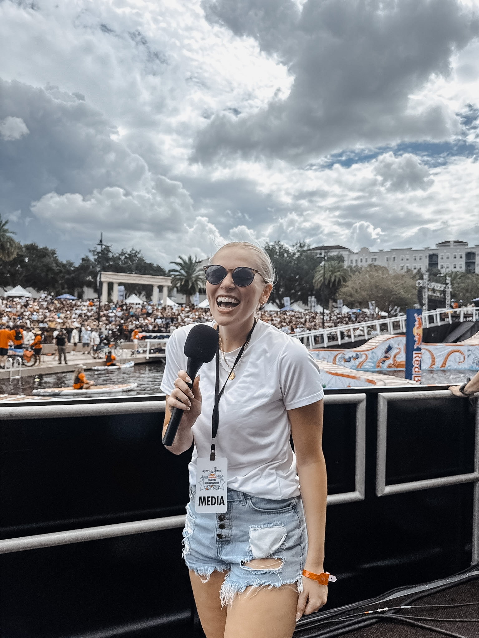 Smiling woman with sunglasses holding a microphone outdoors, crowd and cloudy sky in background.