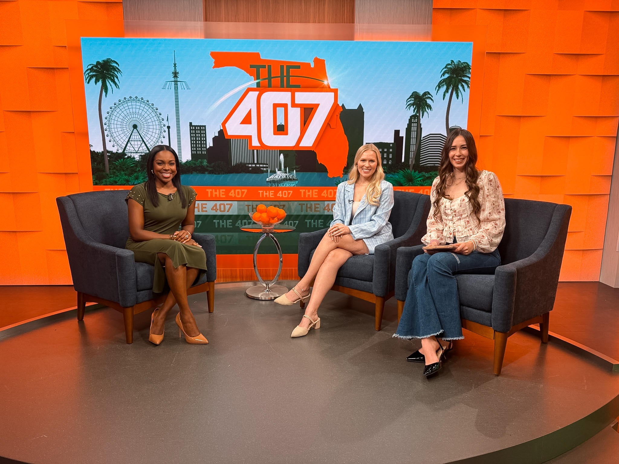 Three women sit on black sofas in a studio with an orange wall and a large screen displaying 'The 407' logo and cityscape.