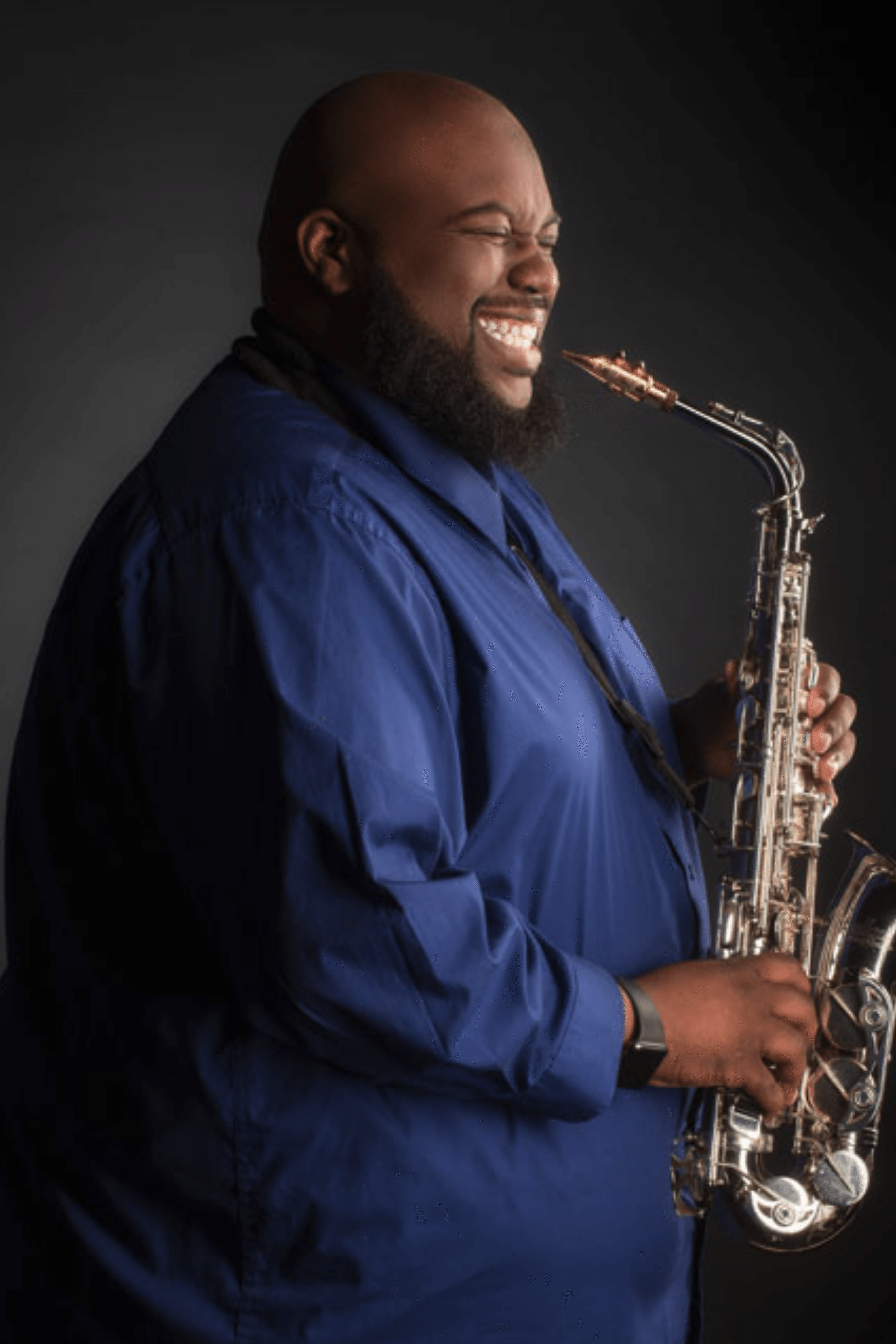 Man playing saxophone, smiling, wearing a blue shirt, against a dark background.