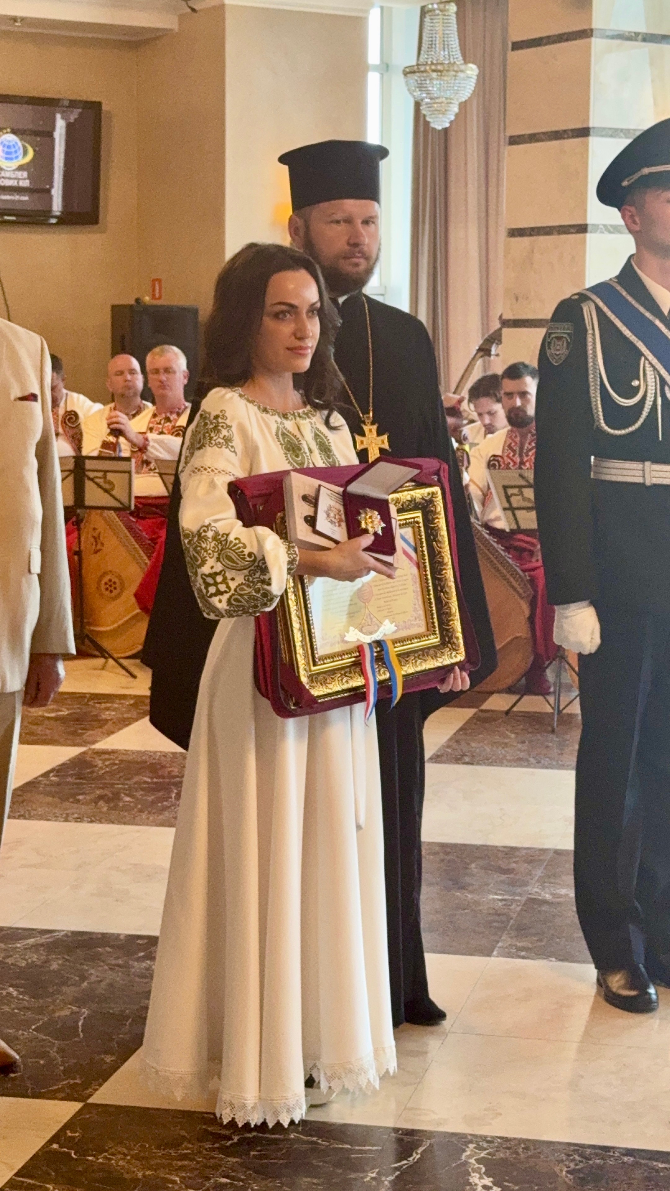 Young woman in white dress holding a framed certificate, standing with two men in traditional attire inside a decorated hall.